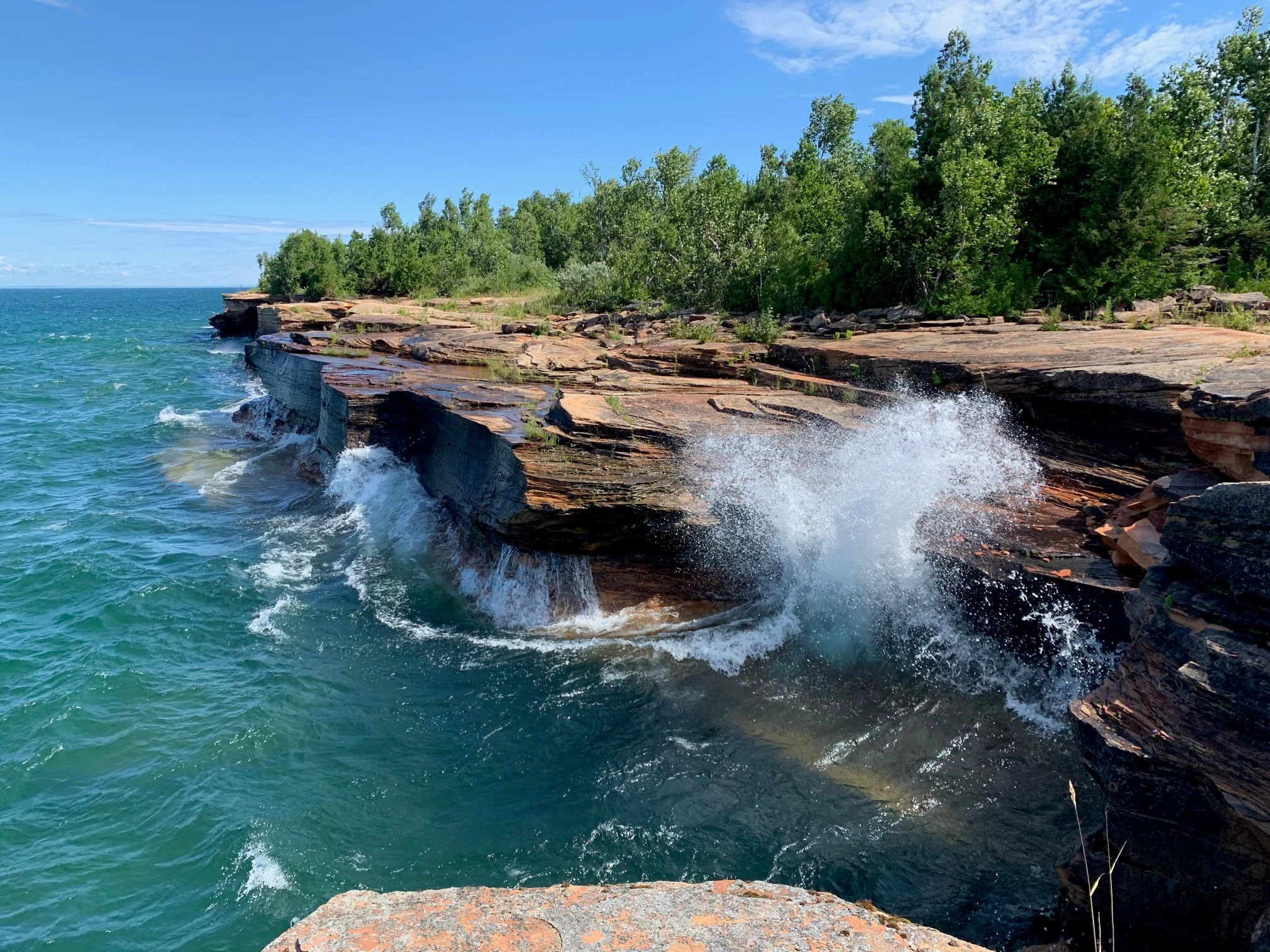 Large white waves crashing against layered red sandstone rock formations along a shoreline, with green trees in the background under a blue sky.