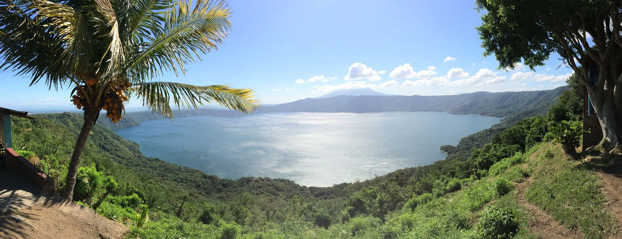 A wide panoramic landscape shot overlooking the large, circular volcanic lake of Laguna de Apoyo, framed by tropical palm and broadleaf trees on the crater slopes.