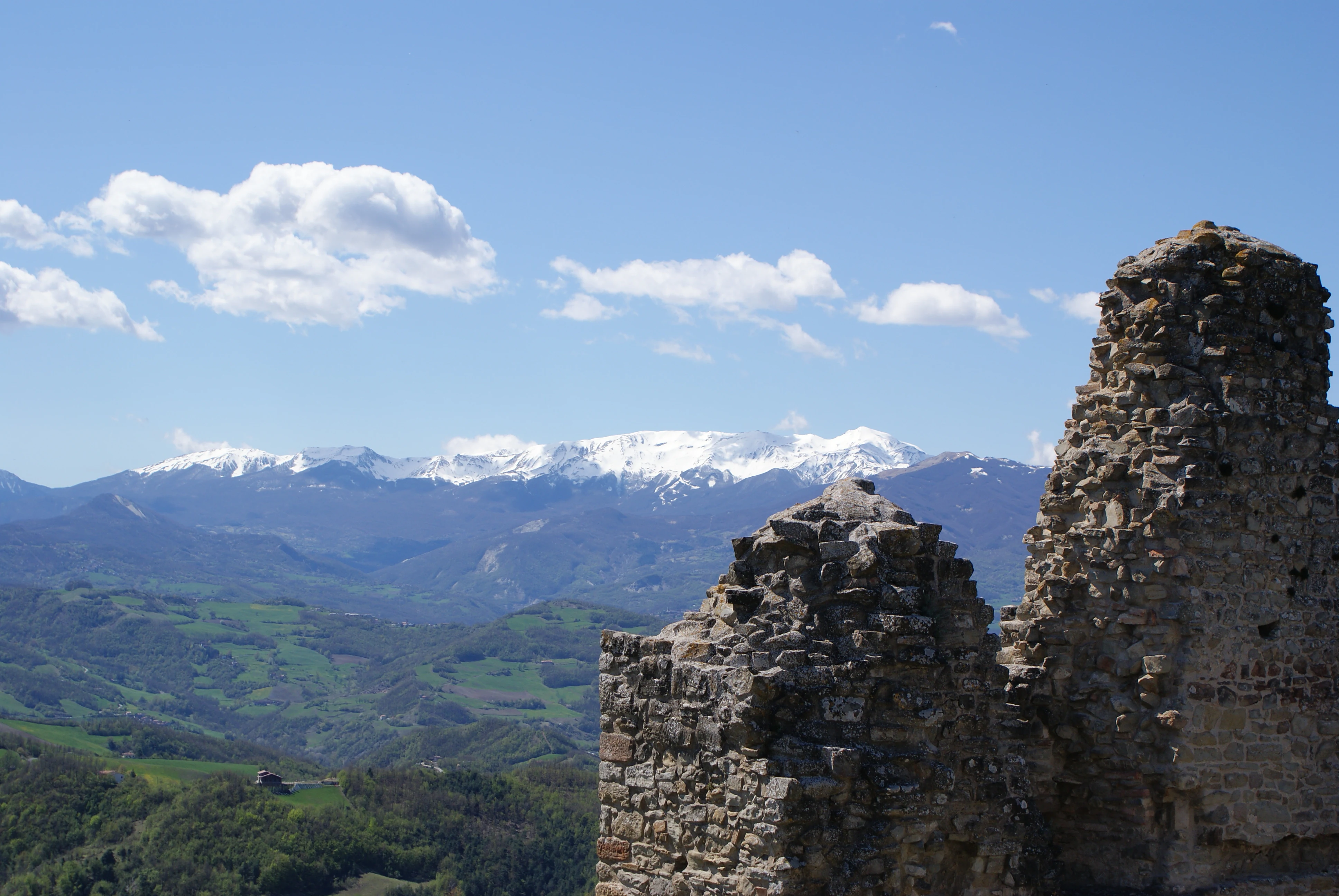 Stone ruins of Carpineti Castle with green valleys and snow-covered mountains under a clear blue sky