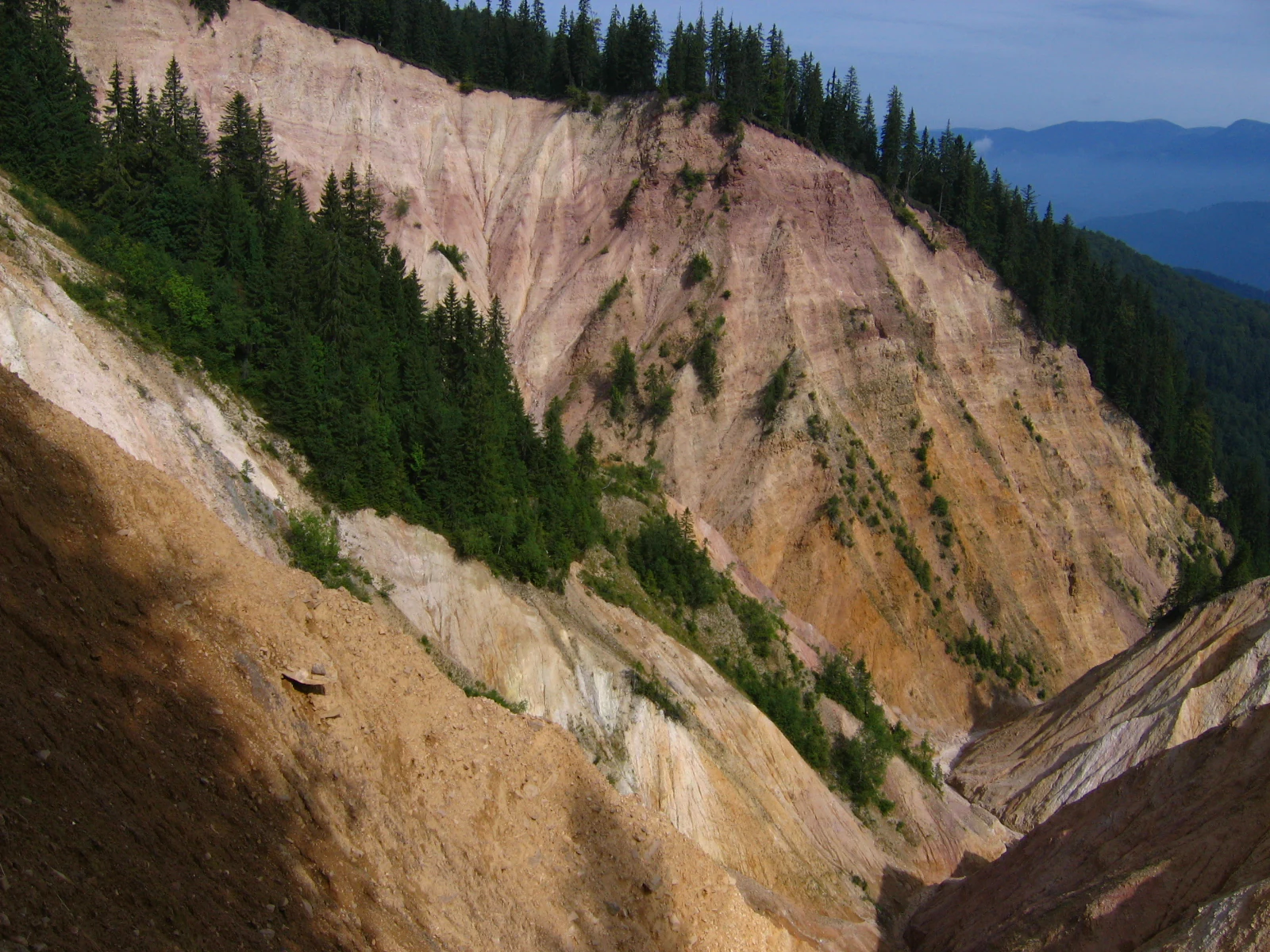 Steep rocky cliff face with exposed earth layers in shades of brown, red, and beige, interspersed with patches of green vegetation, set against forested hills under a partly cloudy sky
