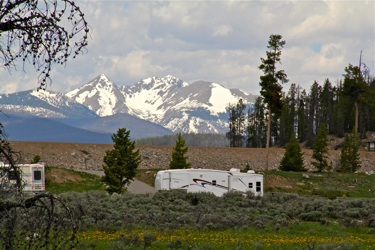 A white recreational vehicle sits in a campground area with sagebrush and pine trees, set against a backdrop of distant, snow-covered mountains.