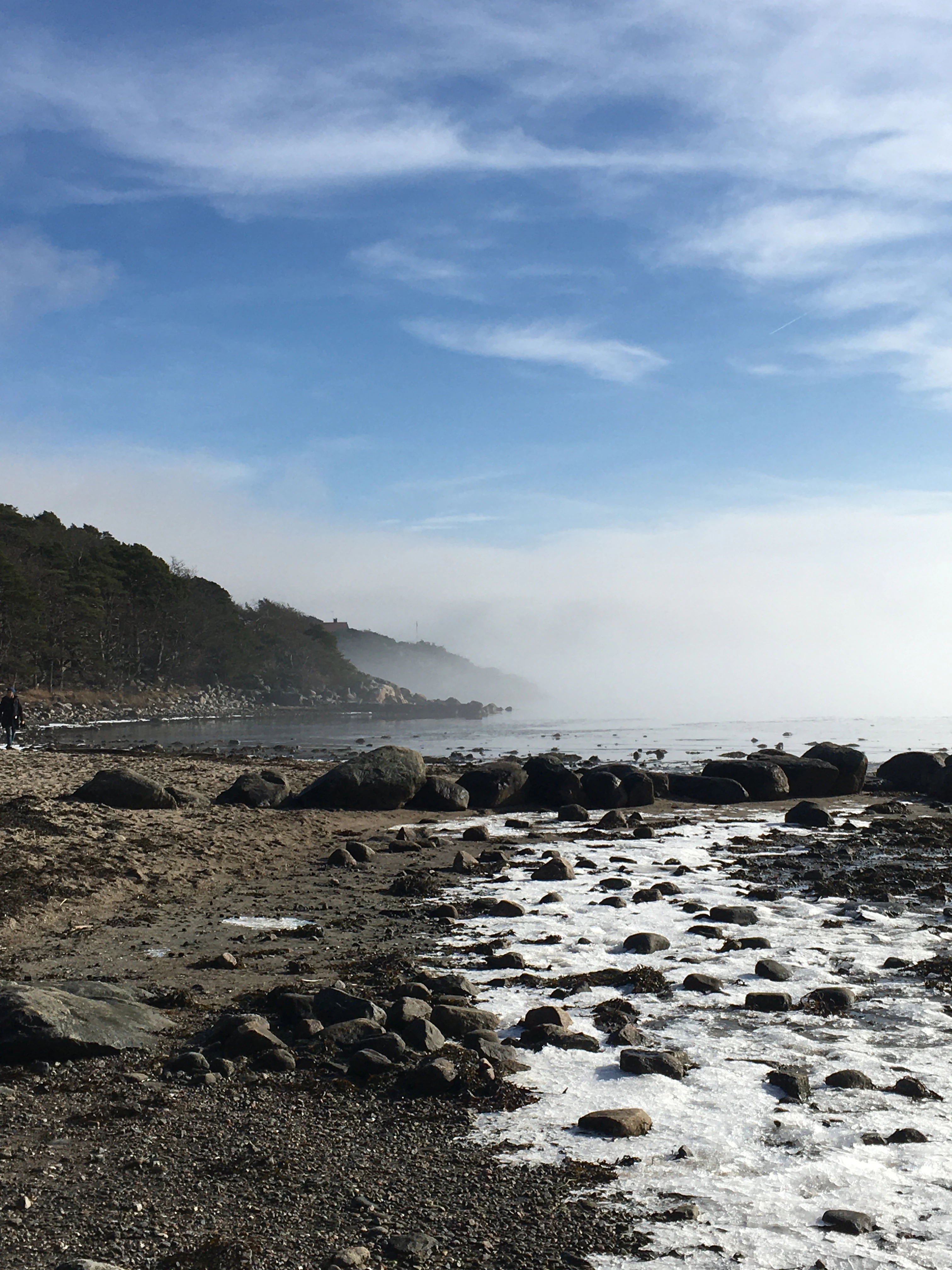 Rocky beach with patches of snow, fog-covered sea, forested hillside, and a person walking under partly cloudy sky