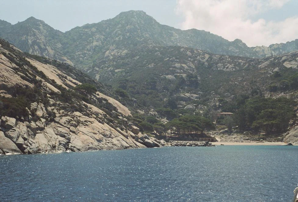Rocky coastline with mountains in the background, calm blue water, and sparse vegetation under a partly cloudy sky