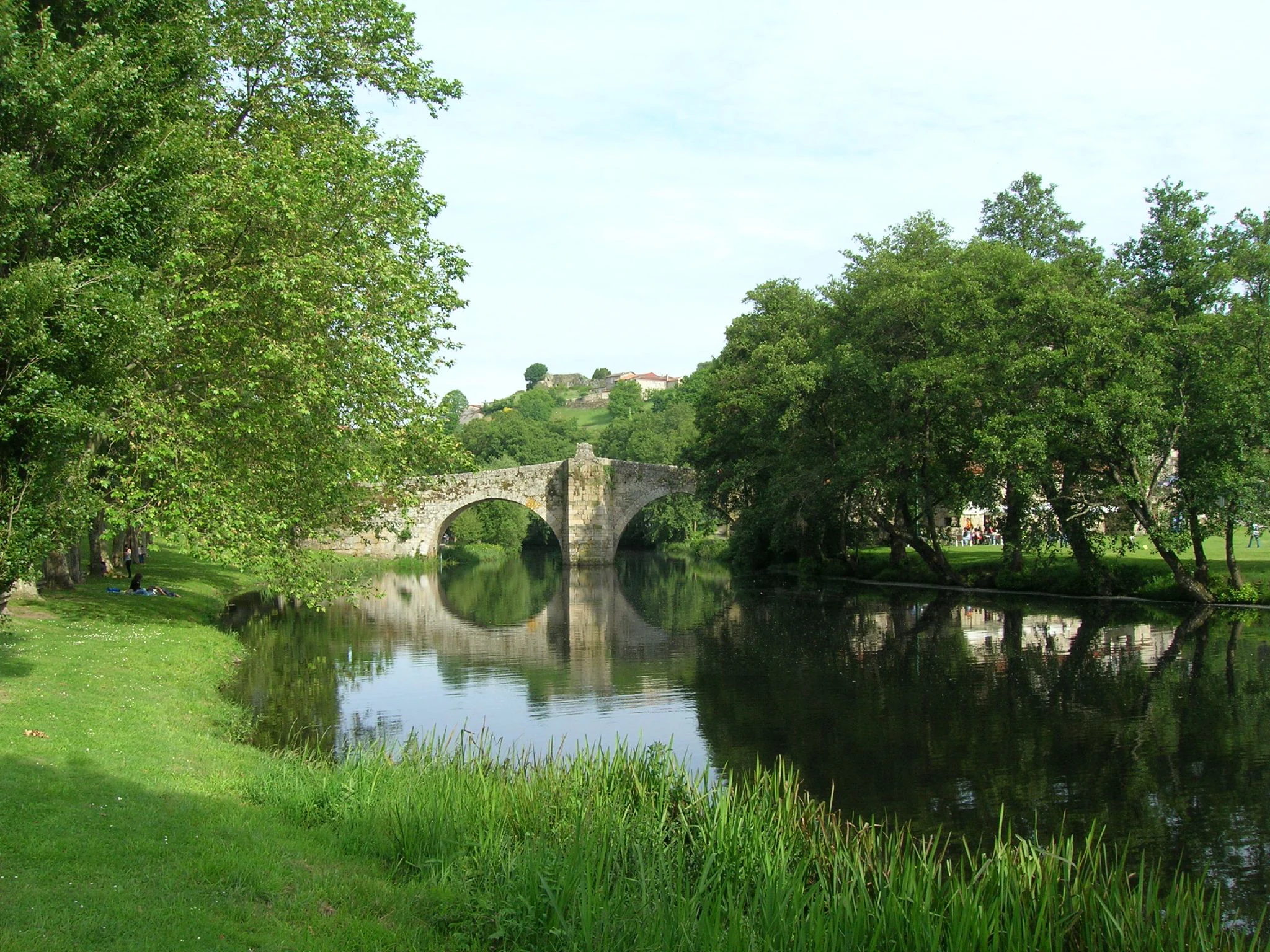A stone arch bridge reflected in the calm waters of the Arnoia River, surrounded by lush green trees and grassy riverbanks.
