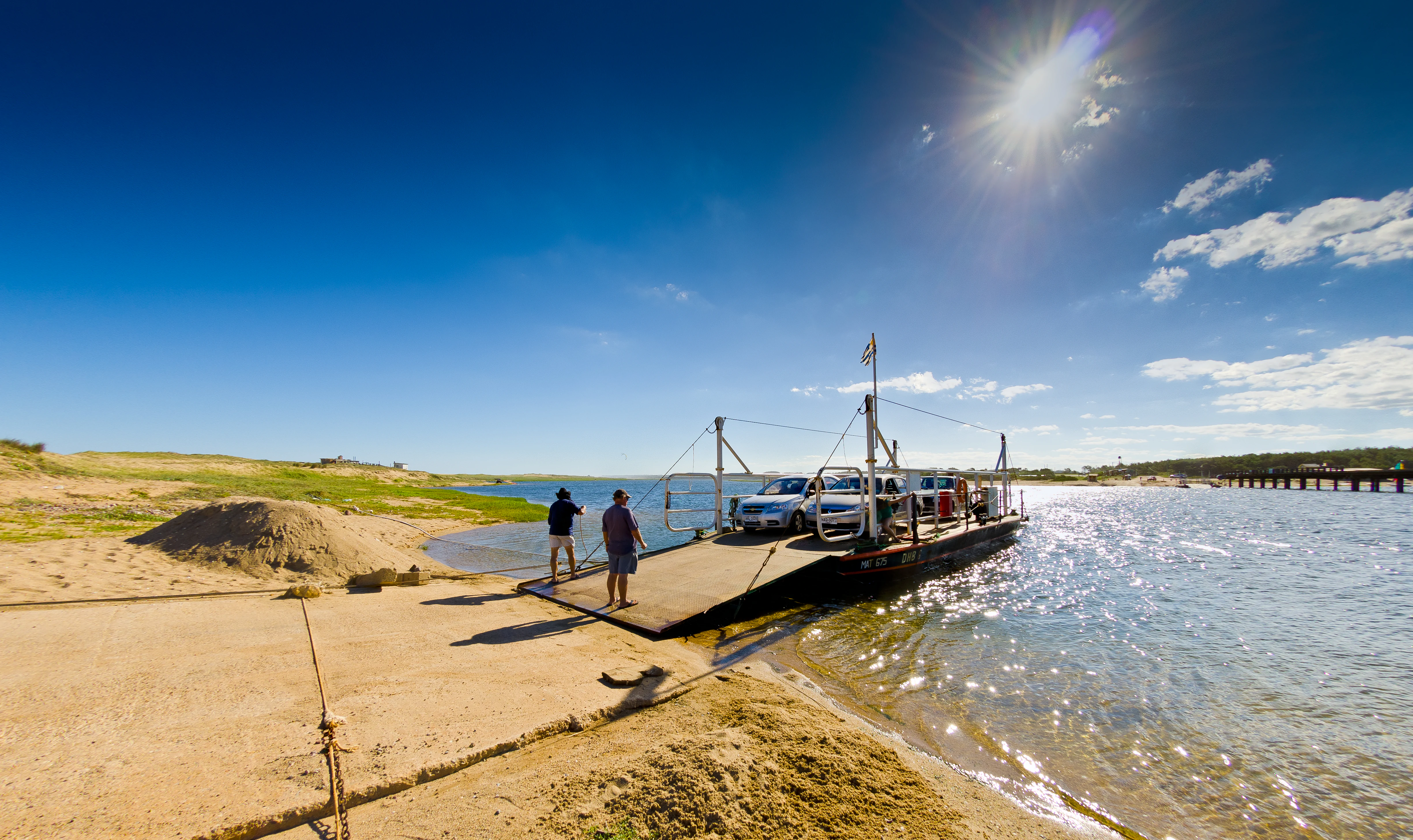 A small flat-deck ferry boat carrying vehicles docked at a sandy shoreline in Laguna Garzon, with two people standing on the ramp in the foreground and bright sunlight overhead.