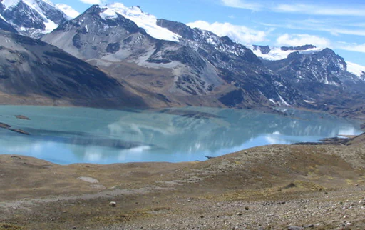 A wide landscape view of a turquoise lake surrounded by rocky terrain and snow-capped mountains under a blue sky.