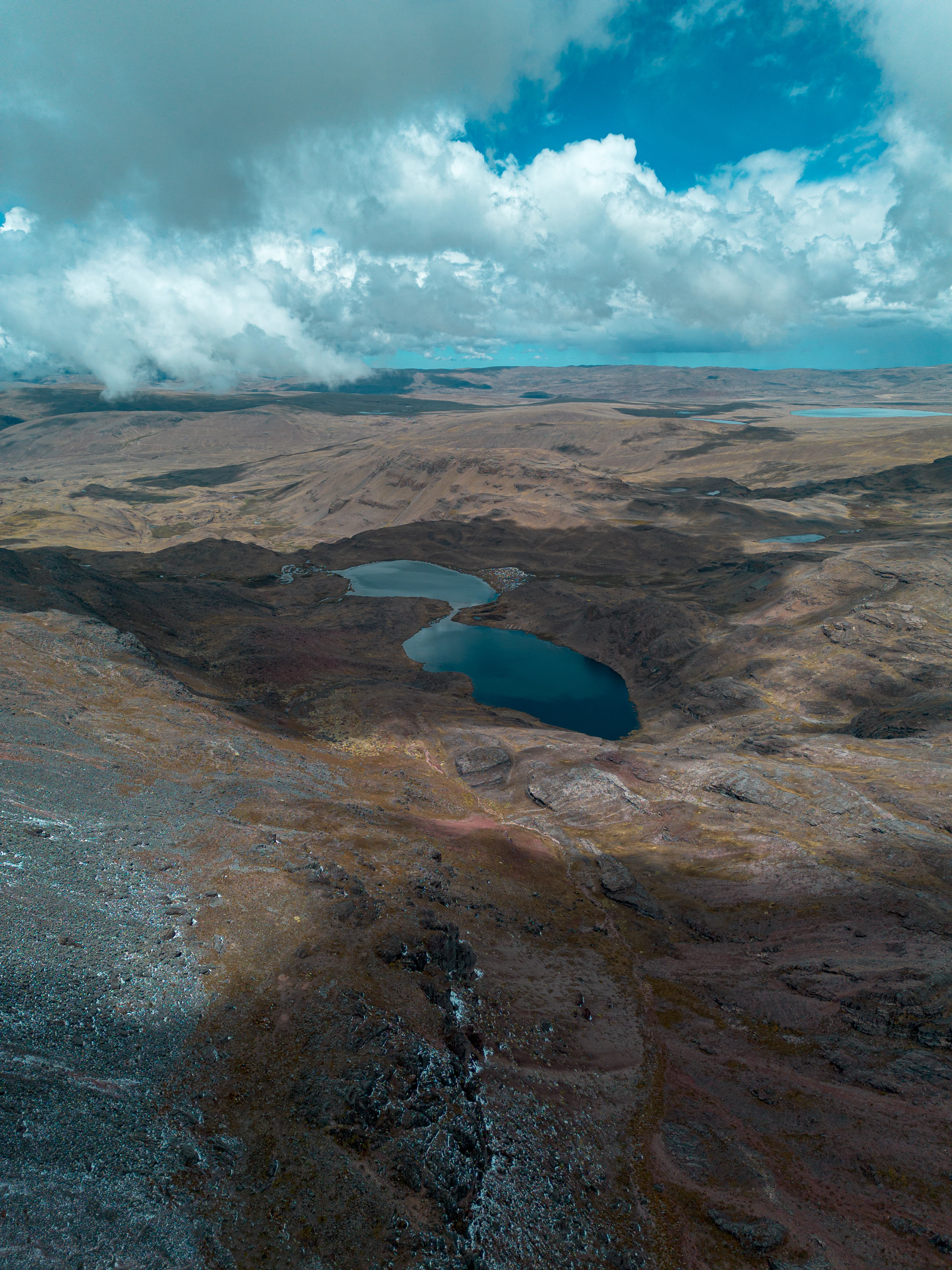 A high angle view of a rugged brownish landscape featuring two small blue lakes nestled in a depression under a partly cloudy sky.