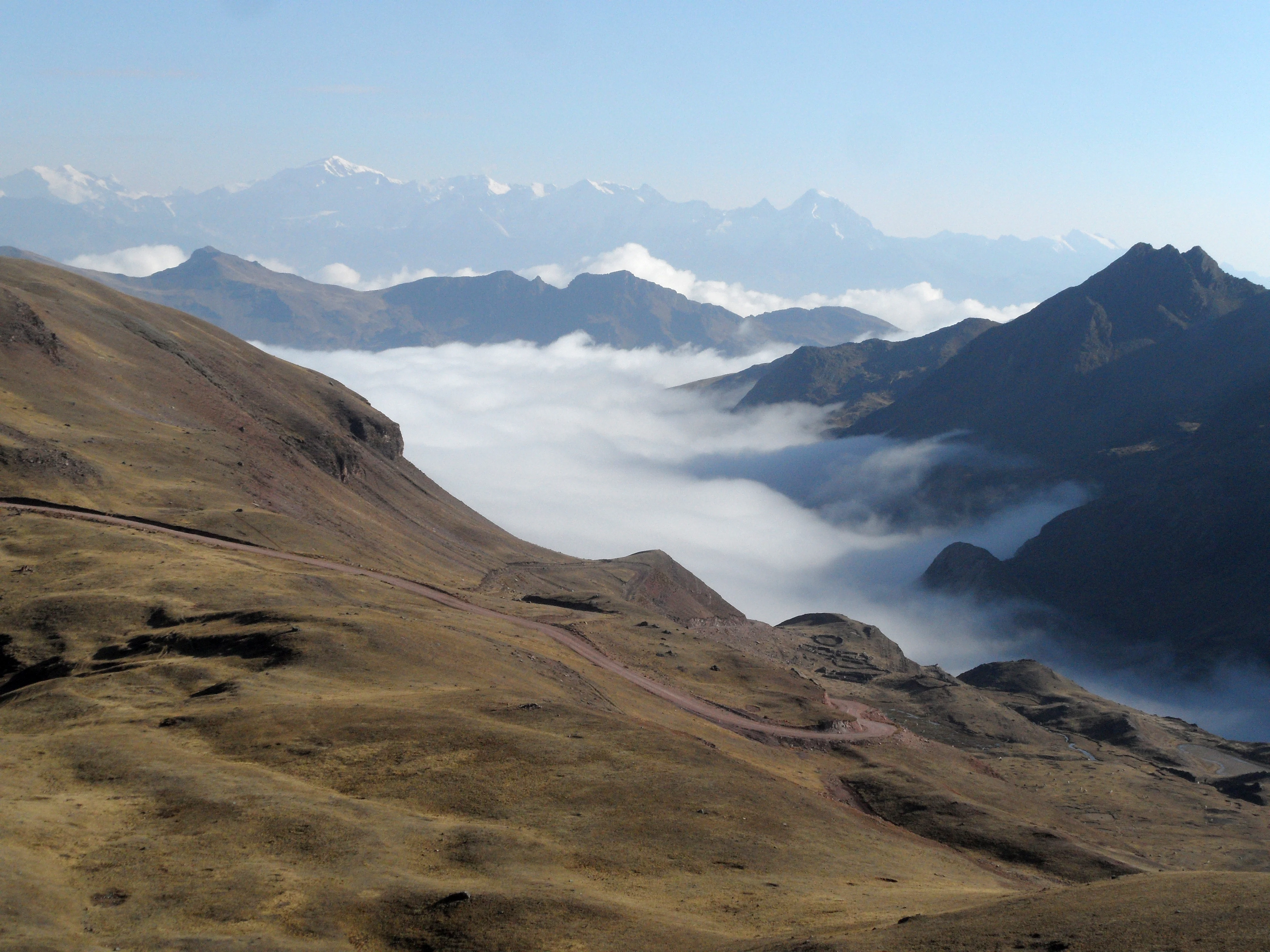 A wide landscape view of high-altitude brownish hills with a winding dirt road, overlooking a valley filled with white clouds and distant snow-capped mountain peaks.