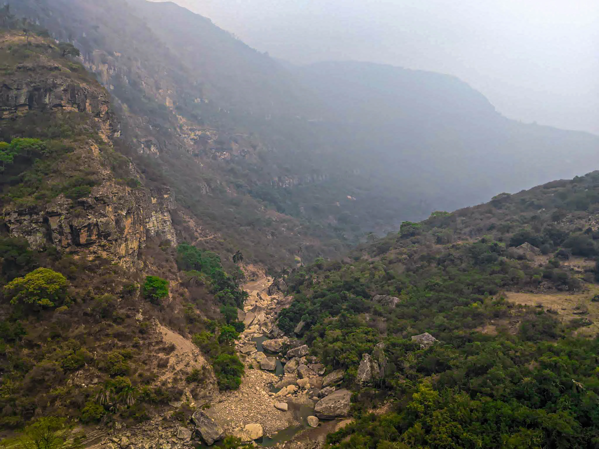 An aerial perspective of a rocky canyon valley featuring steep, vegetation-covered slopes and a dry, boulder-strewn creek bed at the base, under a hazy sky.