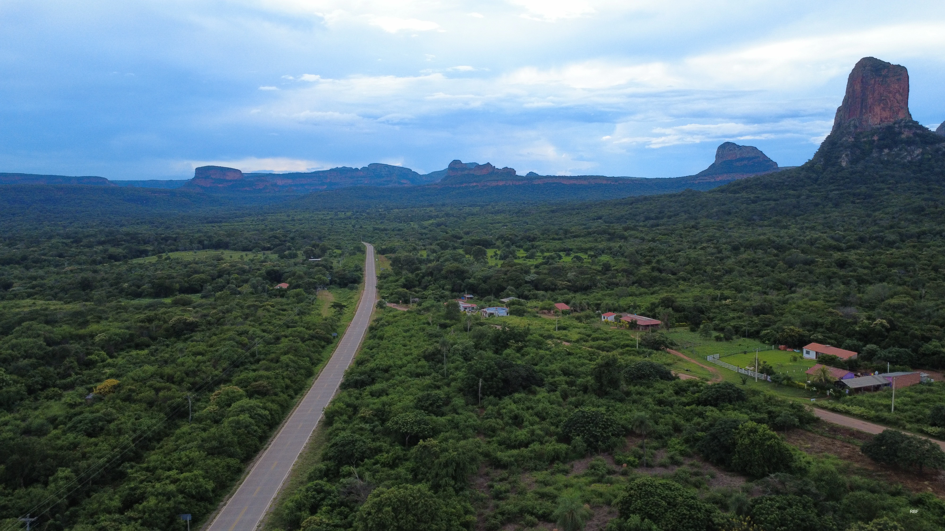 An aerial view of a paved road running through a dense green forest, with prominent, steep red-rock mesas and a tower-like geological formation in the background under a cloudy sky.