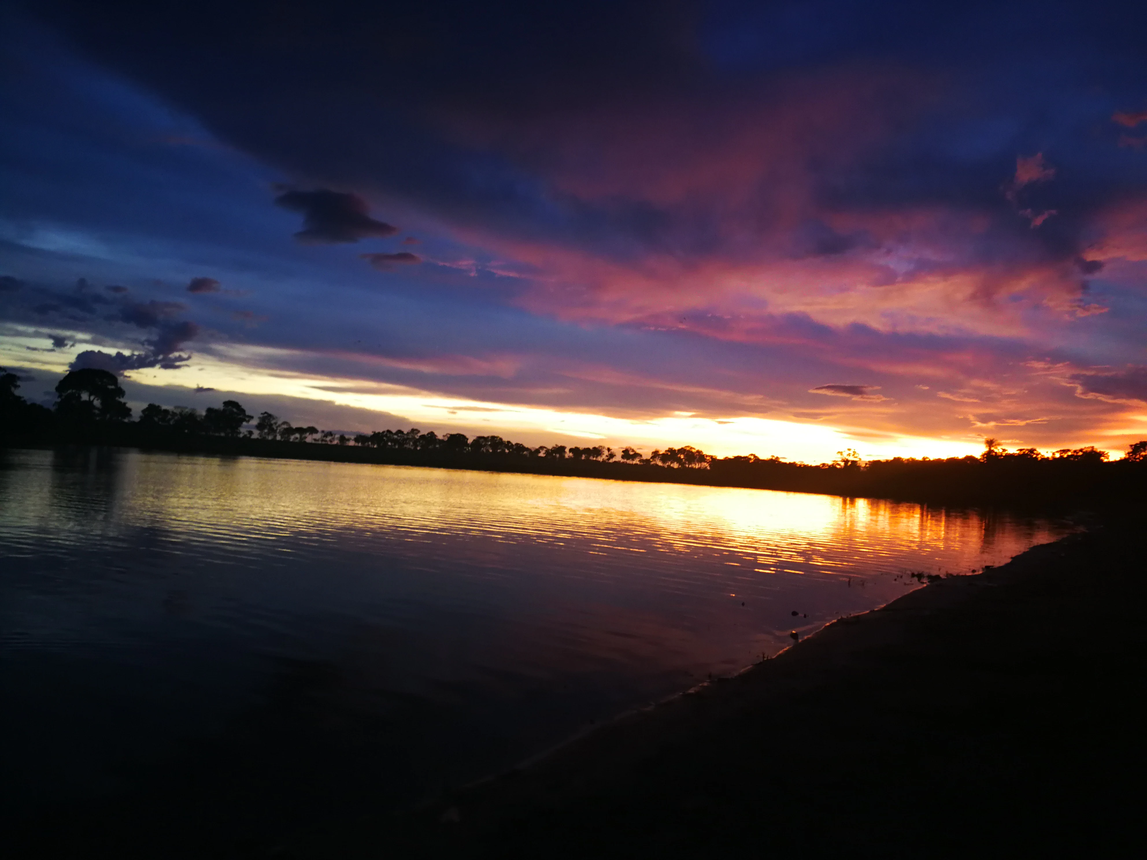 A silhouetted shoreline along a body of water under a vibrant sunset with deep purple, orange, and blue clouds.
