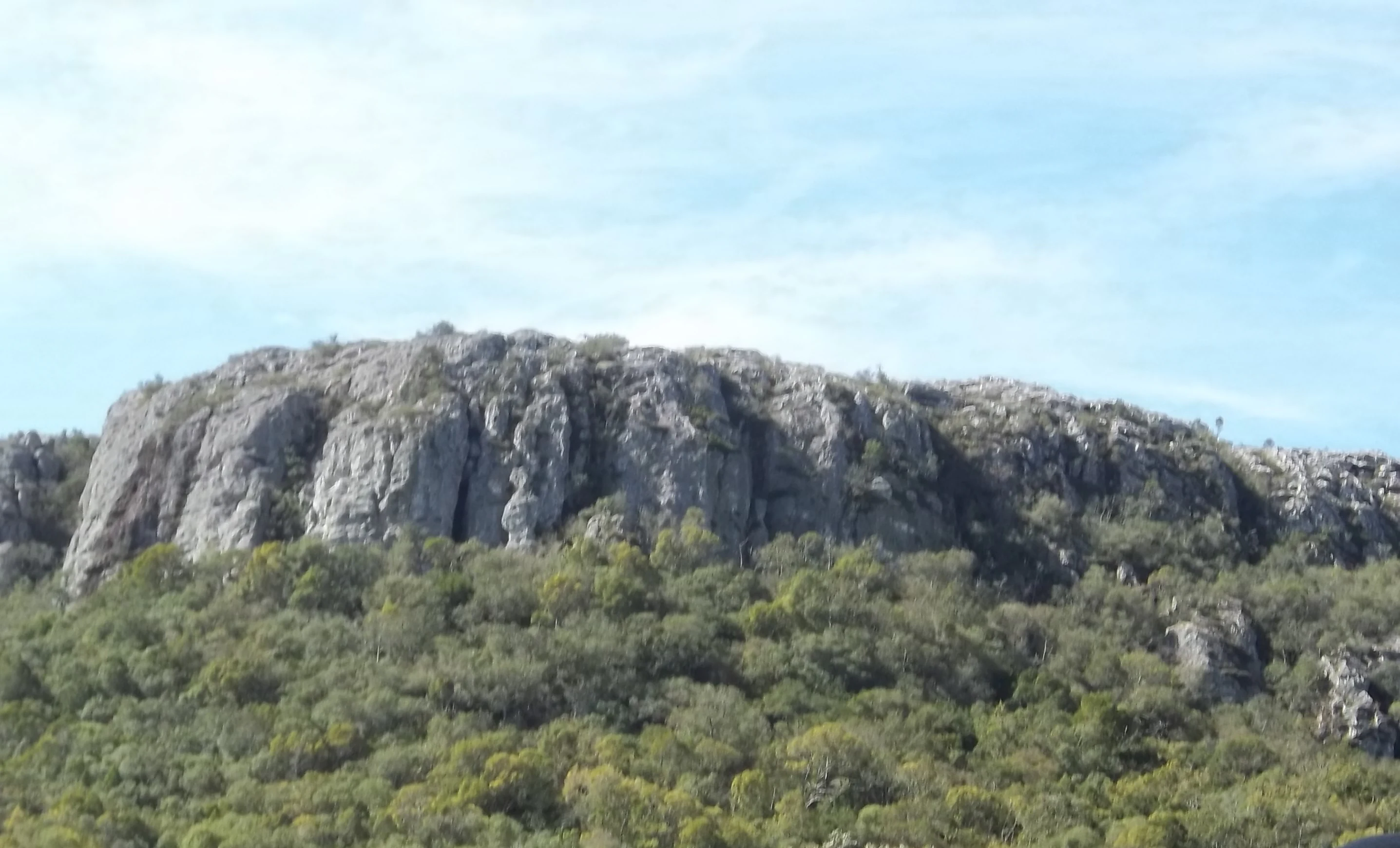 A large rocky hill with a forested base under a partly cloudy sky