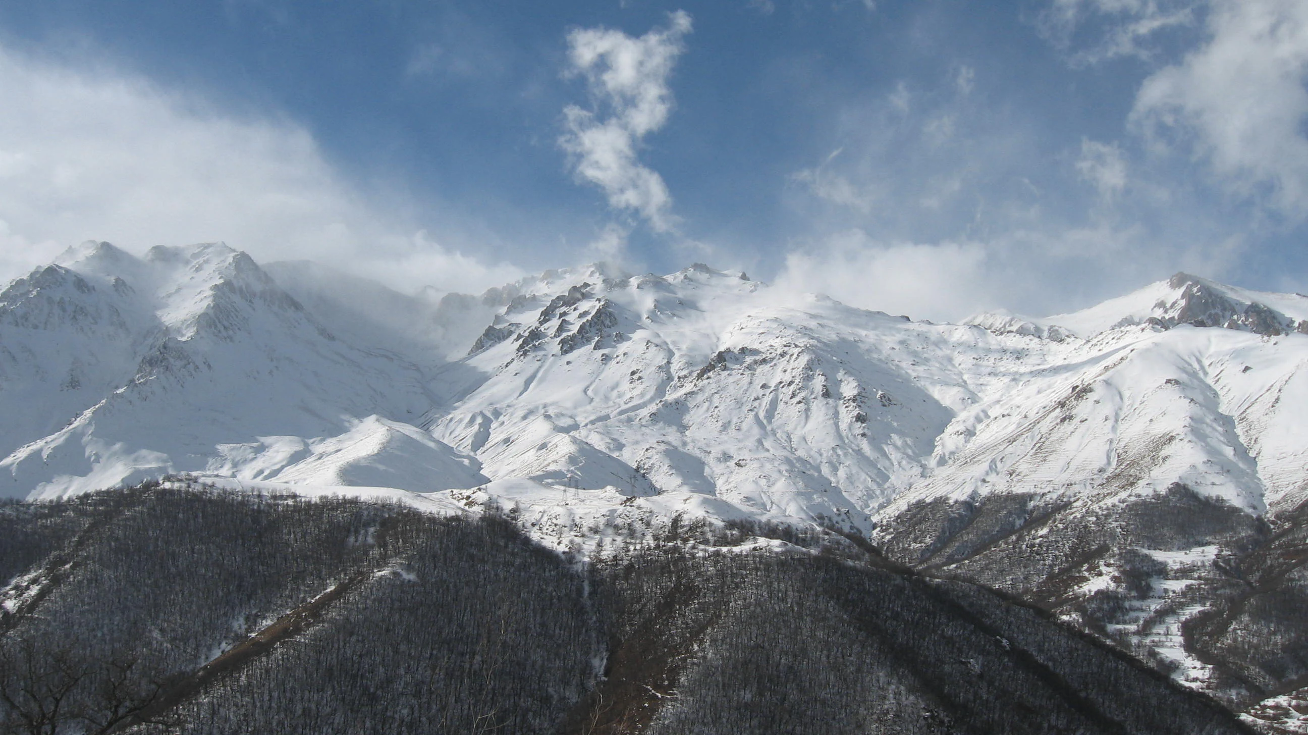 Snow-covered mountain peaks under a partly cloudy sky with forested slopes in the foreground
