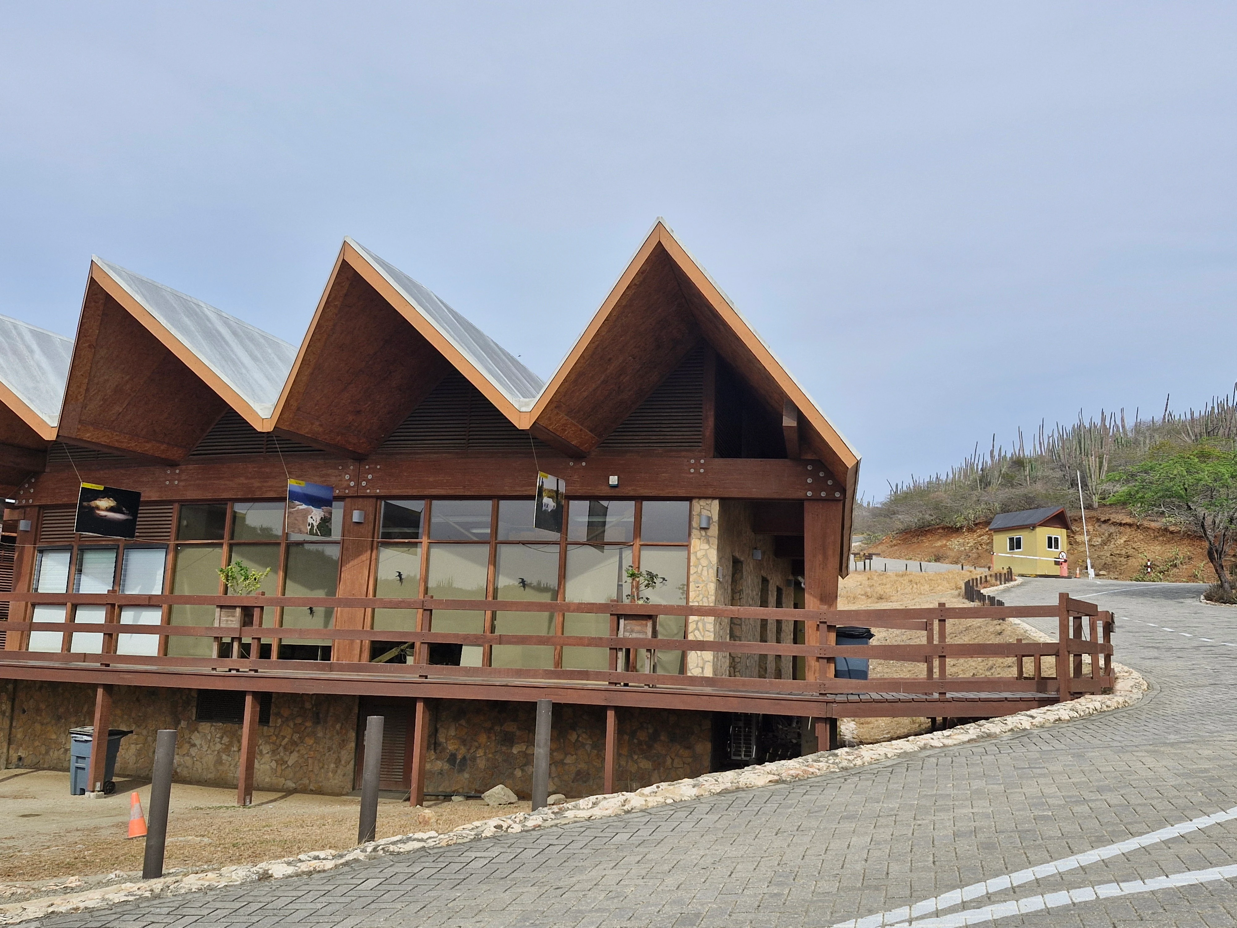 A modern building with a multi peaked, angular wooden roof serves as the visitor center at Arikok National Park in Aruba, with a paved road leading past it.