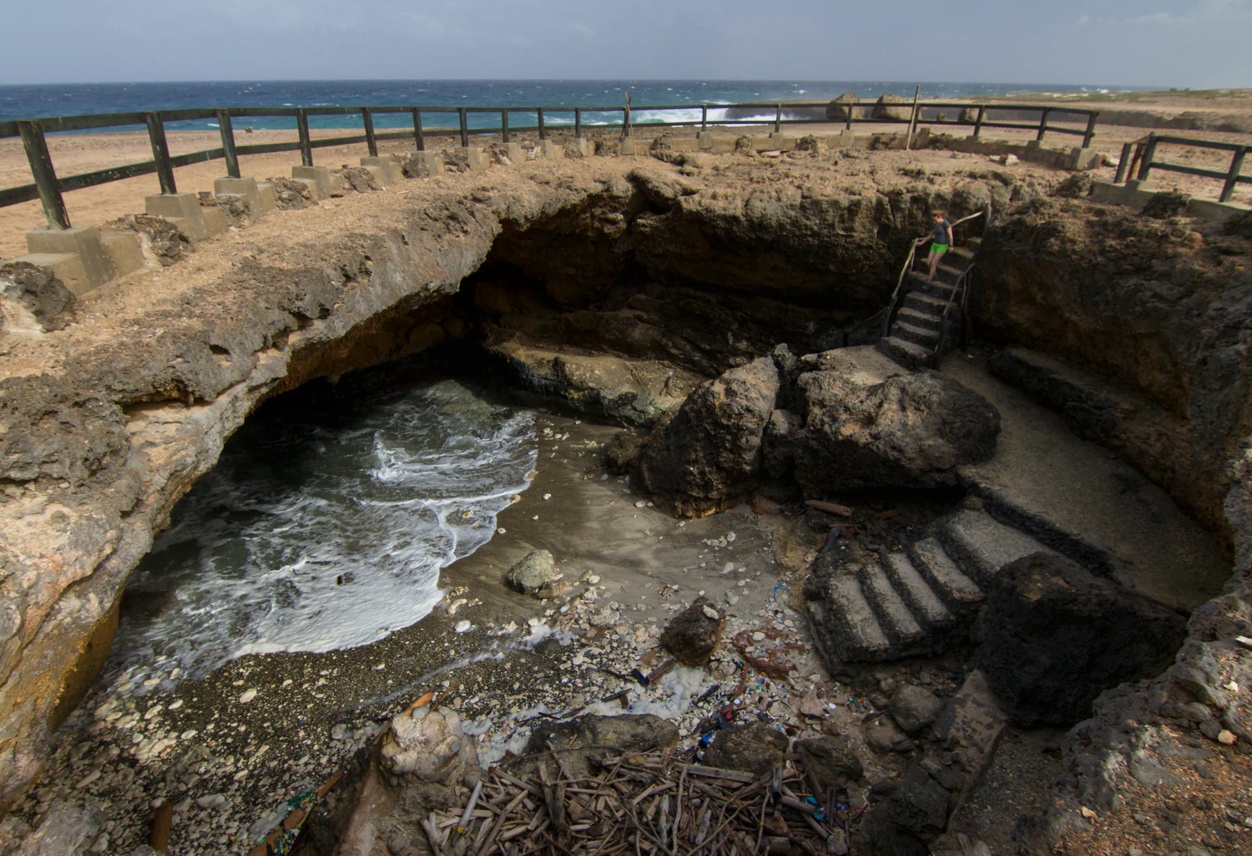 A wide angle shot of a large limestone sinkhole at the coast, with stairs leading down to a small sandy beach where waves enter a sea cave, protected by a wooden railing above.