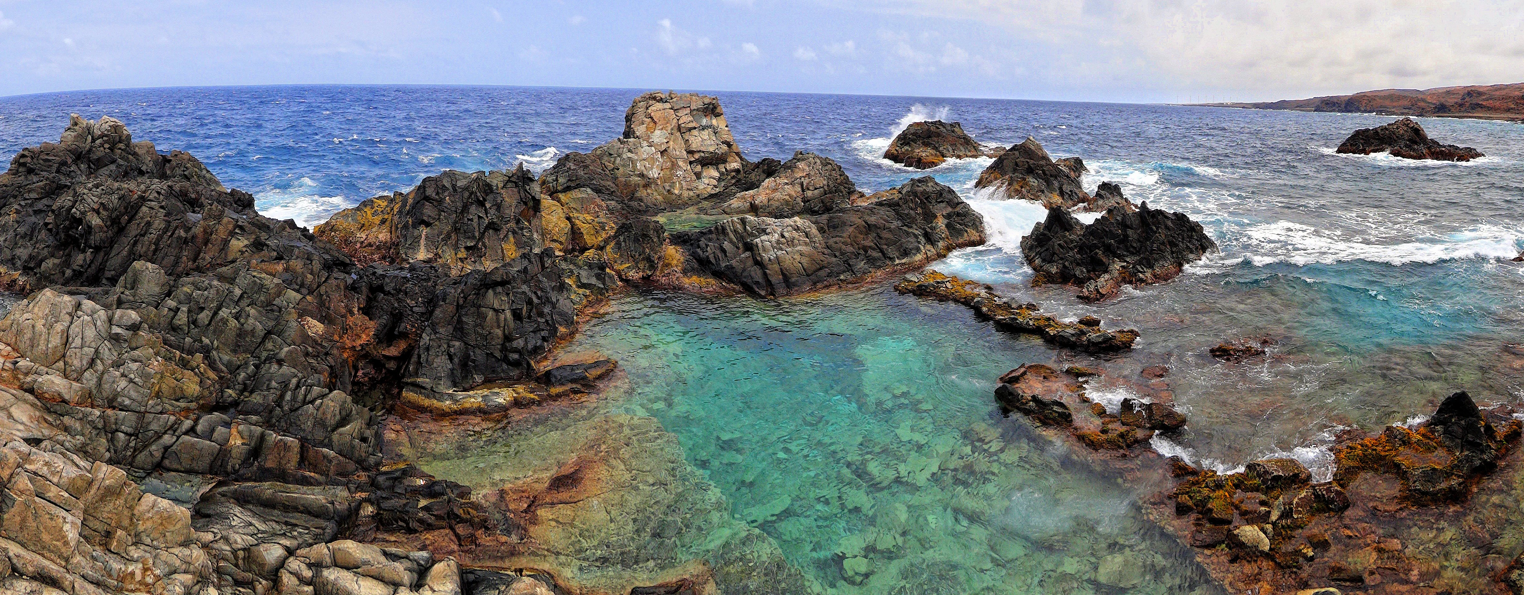 A panoramic view of the Conchi natural pool in Arikok National Park, featuring clear turquoise water contained by dark, rugged volcanic rock formations surrounded by the rough ocean.