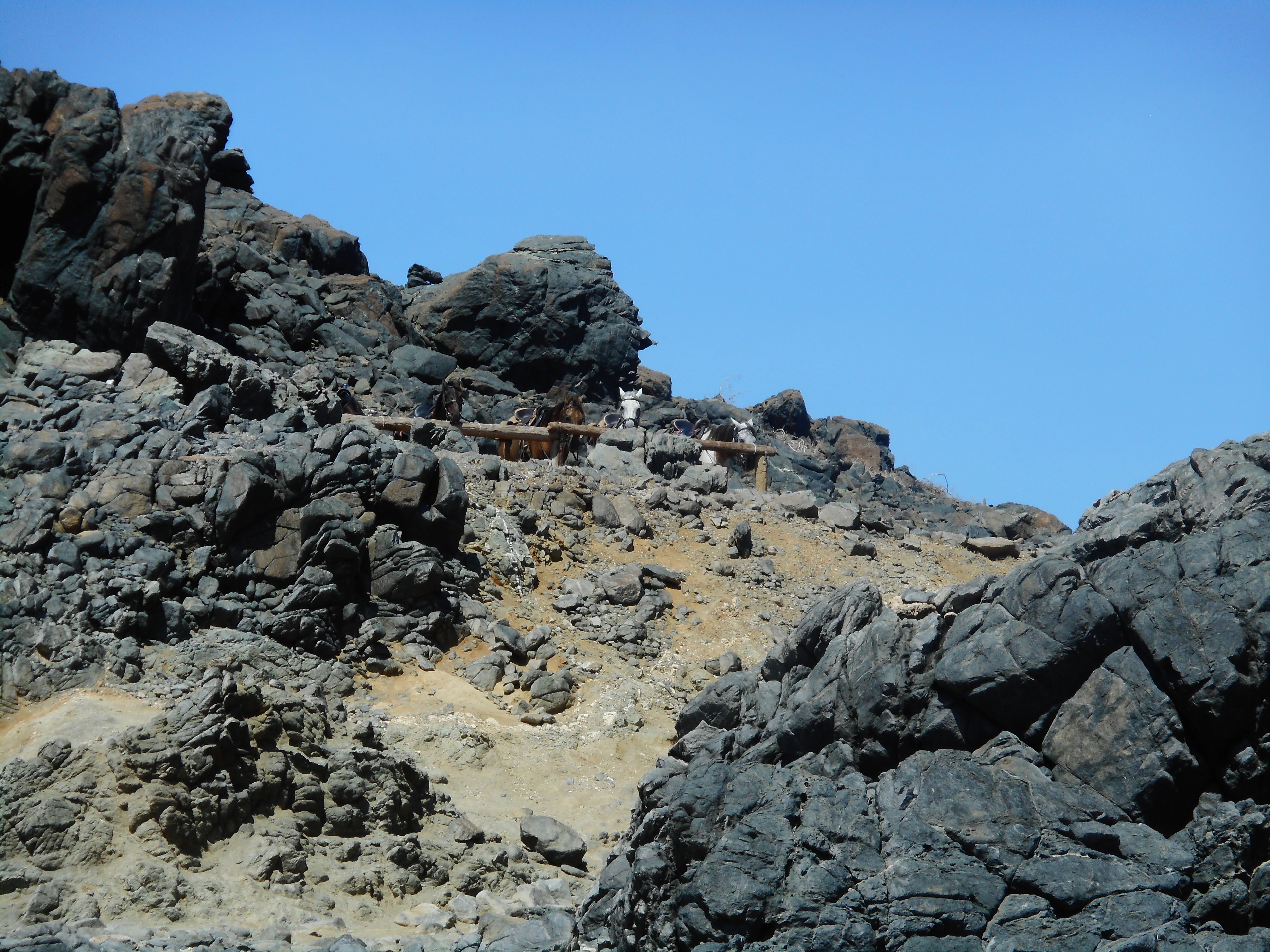 A rocky, arid landscape with dark volcanic lava rocks and lighter, sandy ground under a clear blue sky.