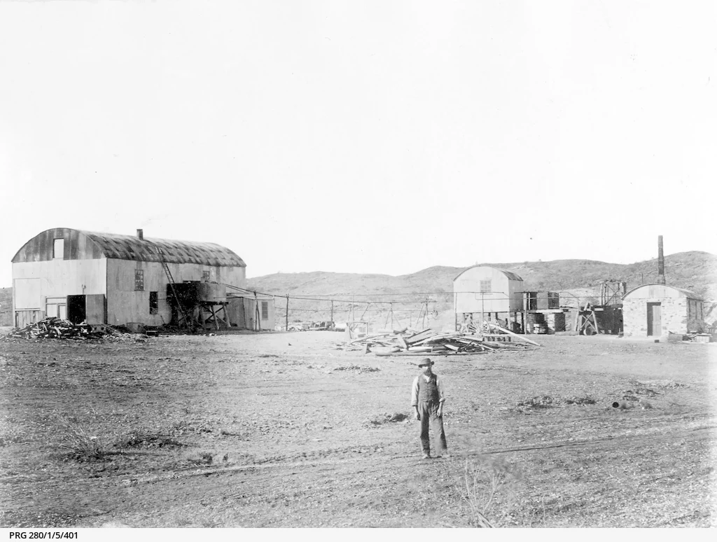 A black and white historical photograph showing several corrugated iron buildings in an arid, rocky landscape. A lone person stands in the foreground, with distant hills in the background.