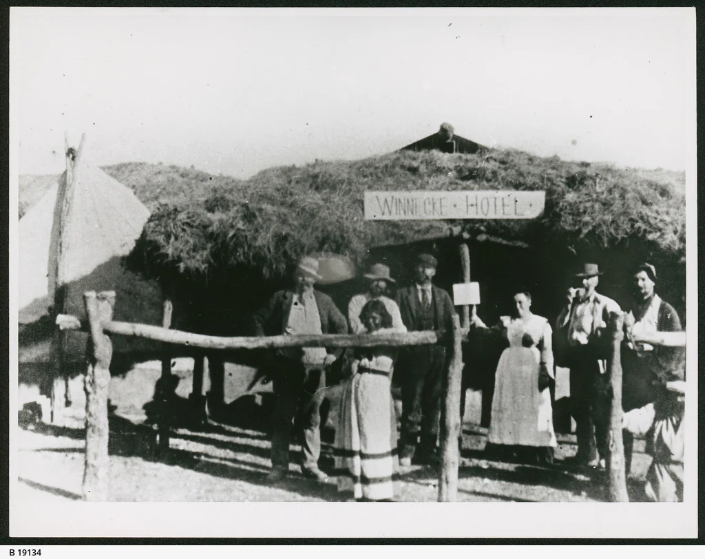 A black and white archival photograph showing a group of people standing in front of a rustic building with a thatched roof, identified by a sign reading Winnecke Hotel.