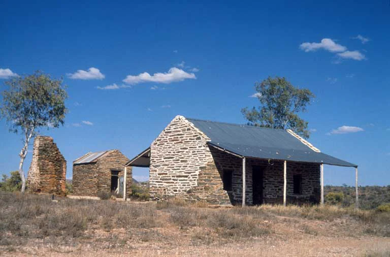 A view of two historic stone buildings under a bright blue sky at Arltunga Historical Reserve, featuring dry desert brush and sparse trees.