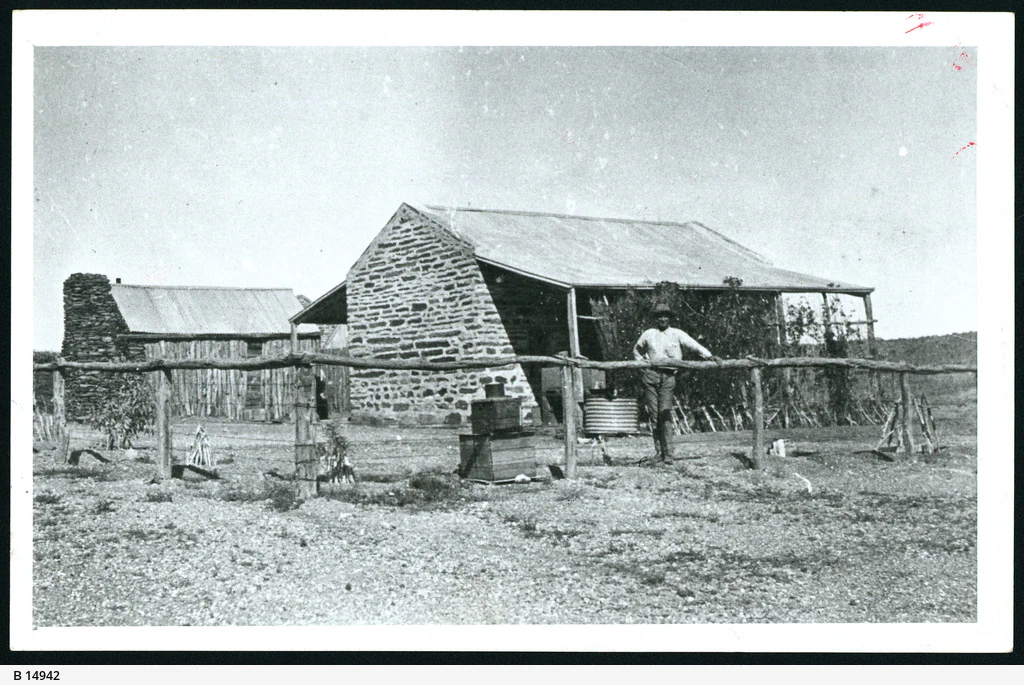 A black and white historical photograph showing a man standing in front of a stone police station building in Arltunga, with a simple wooden fence in the foreground.