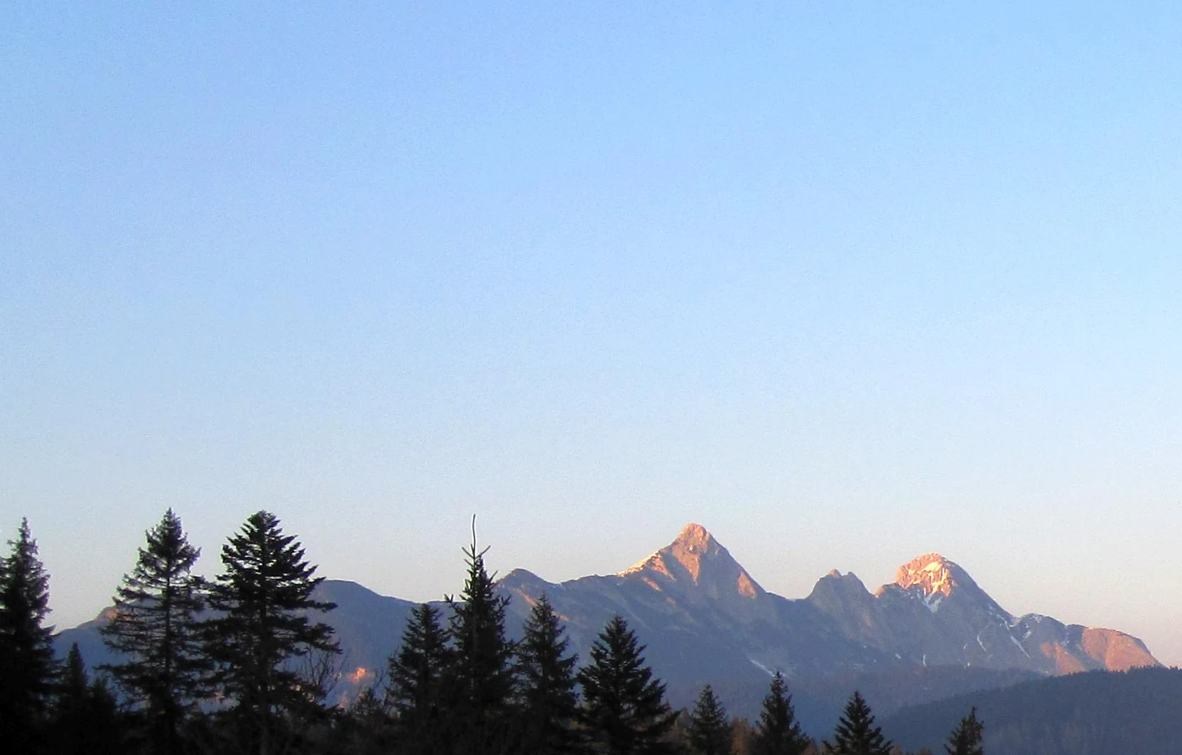 A mountain range with jagged peaks silhouetted against a pale blue sky, with a line of coniferous trees in the foreground.