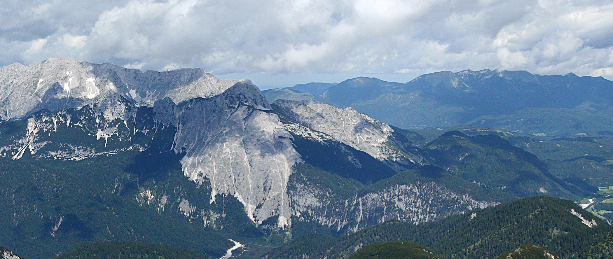 A wide-angle landscape shot showing a rugged limestone mountain range under a cloudy sky, with pine forests covering the lower slopes and valleys below.