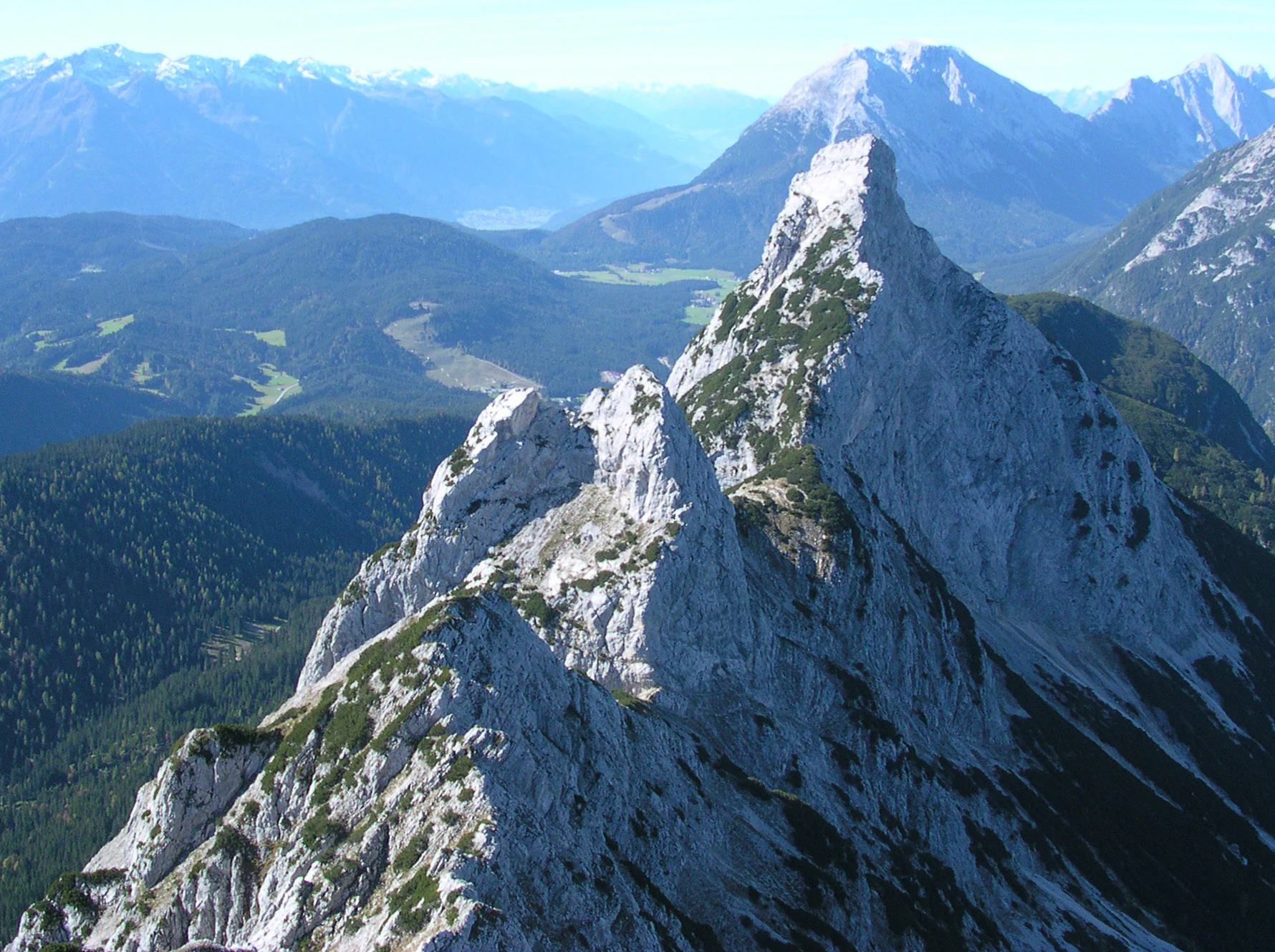 A high-angle, eye-level view of rugged, light-colored limestone peaks in the Arnspitze Group, with forested valleys and distant mountain ranges under a clear sky.