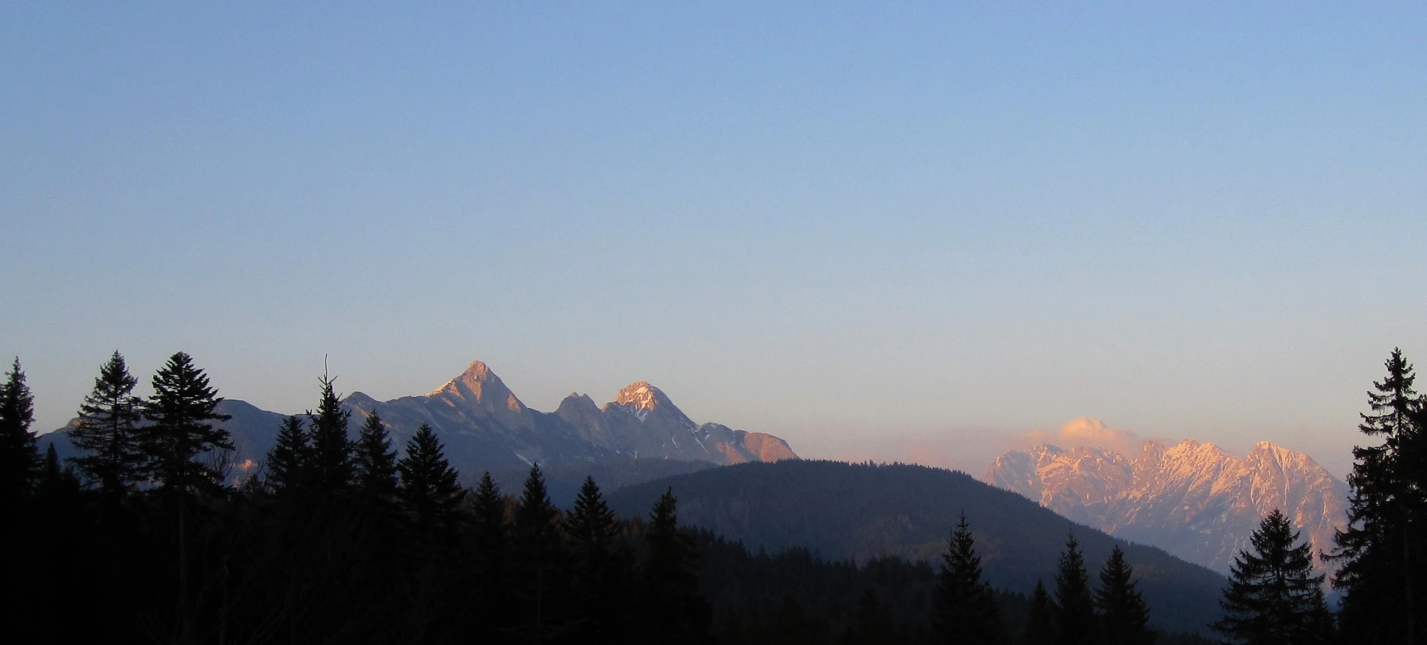 A wide, scenic view of jagged, pale limestone mountain peaks during sunrise, with dark silhouetted pine trees in the foreground.