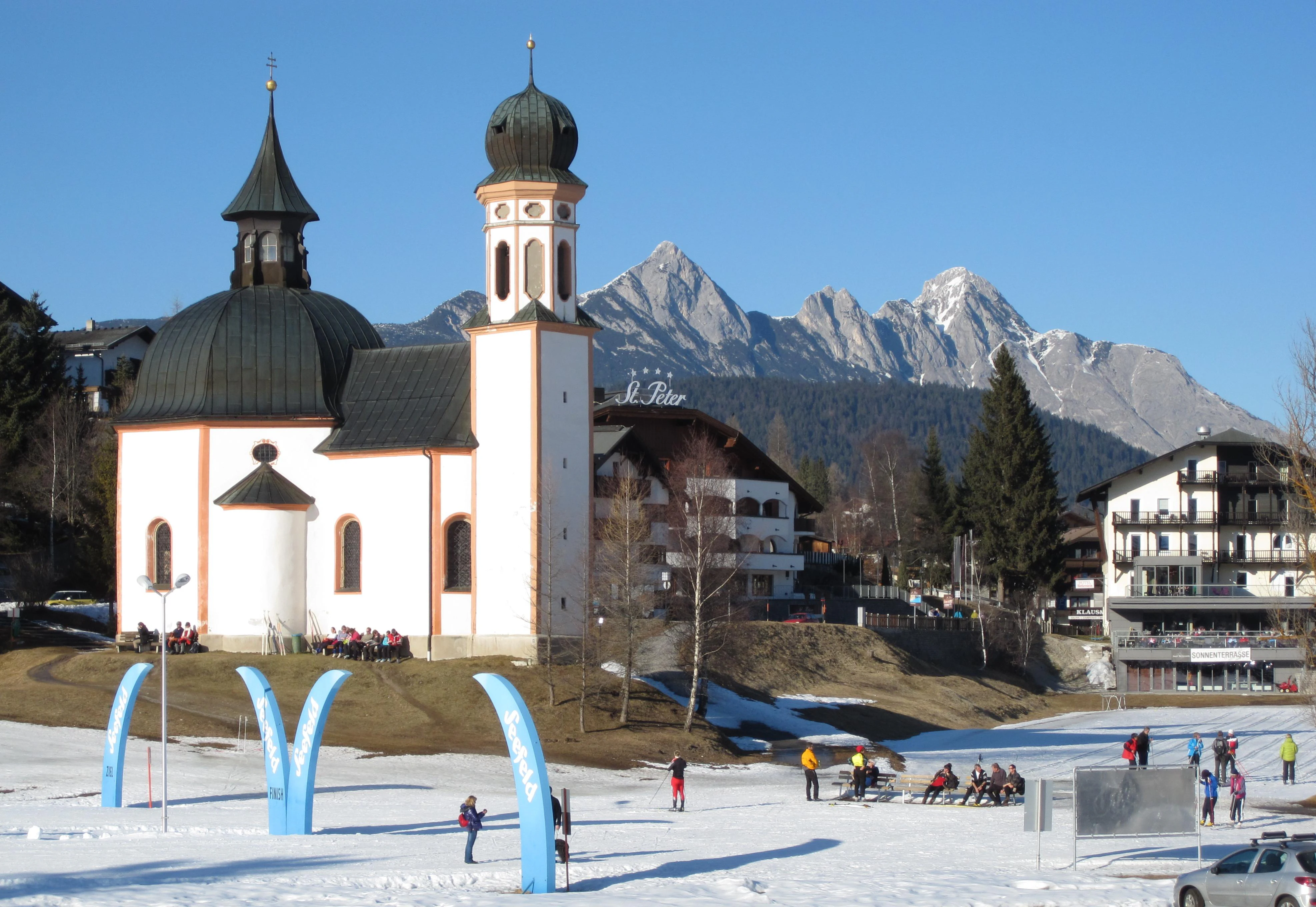 A white chapel with a black dome and tower stands in the foreground with snow on the ground, while a rugged mountain range rises sharply in the background under a clear blue sky.
