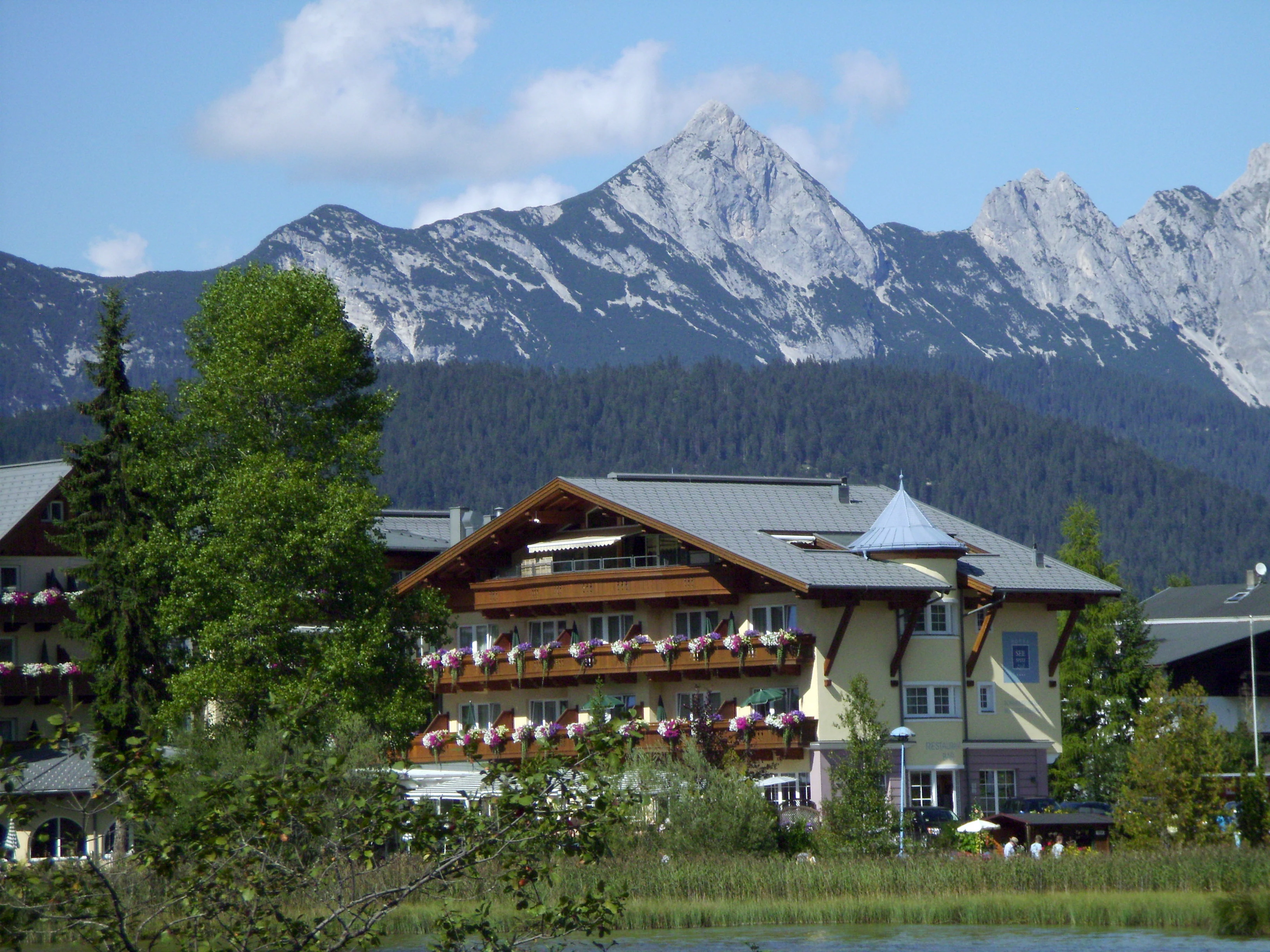 A view of the jagged limestone Arnplattenspitze mountain peak rising behind a traditional Alpine hotel and tree-lined lake shore.