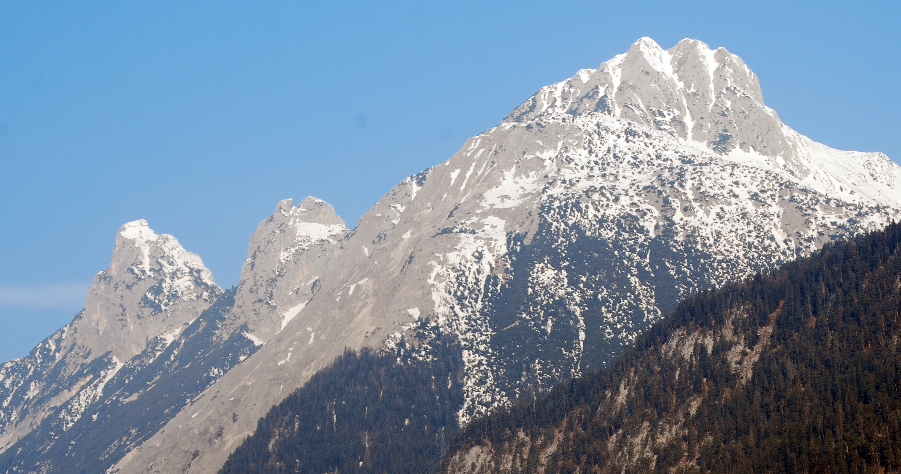 A landscape view of the jagged limestone peaks of the Arnspitze Group with patches of snow under a clear blue sky, featuring foreground forested slopes.