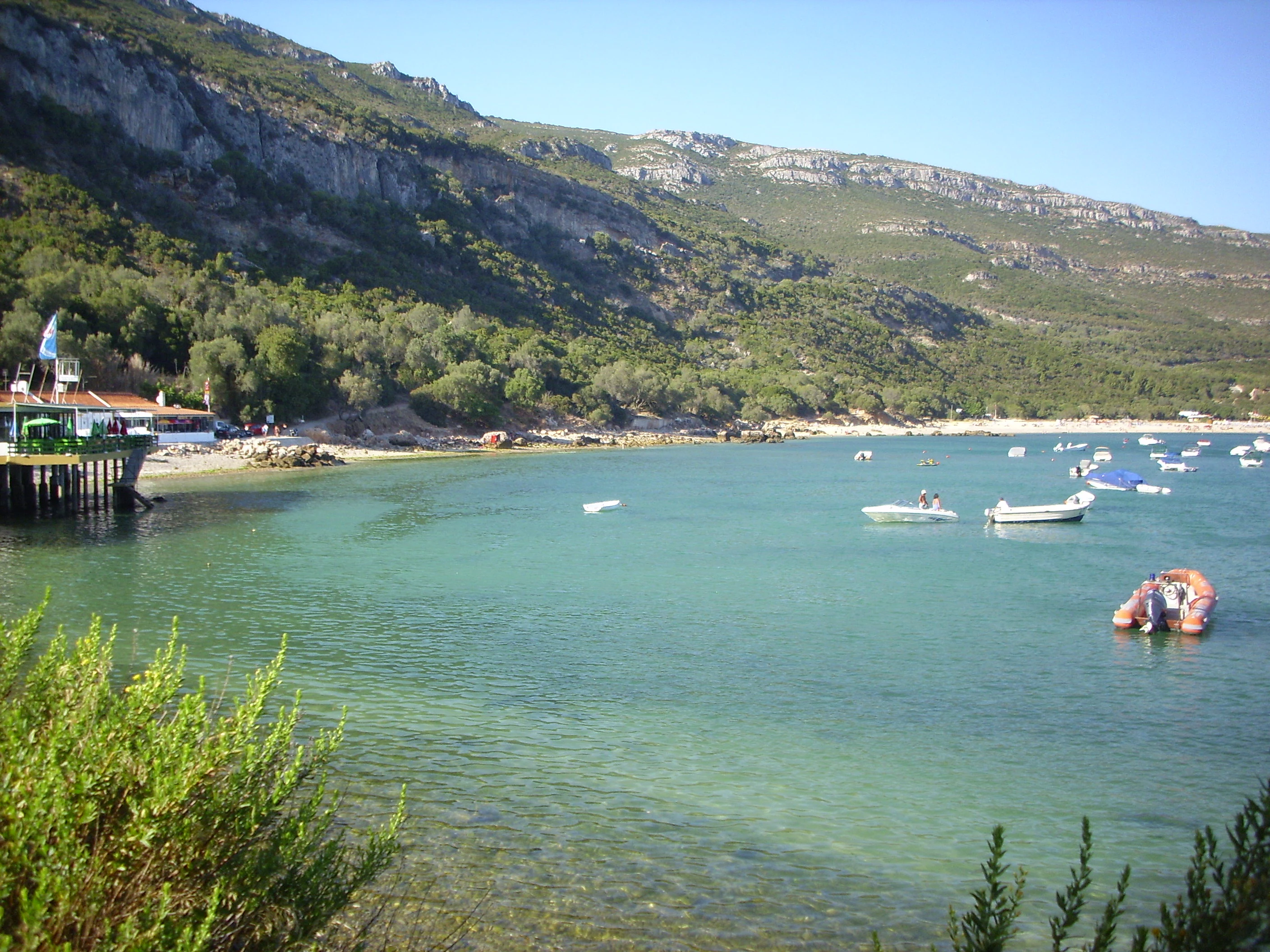 A coastal scene featuring turquoise water, small boats anchored near a sandy beach, rocky cliffs in the background, and a wooden pier on the left side