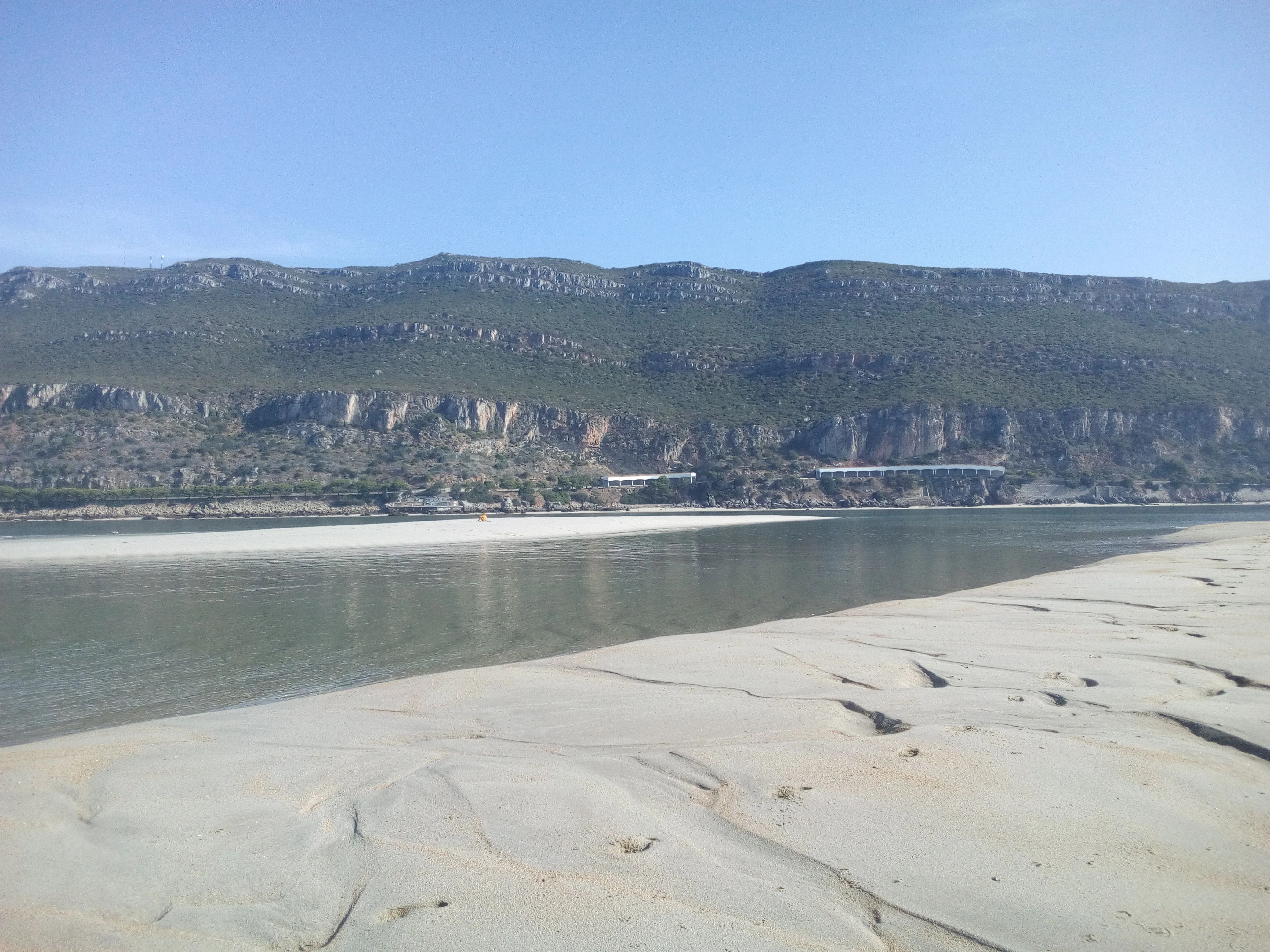 Sandy beach extending into calm water with mountain range in background under clear blue sky