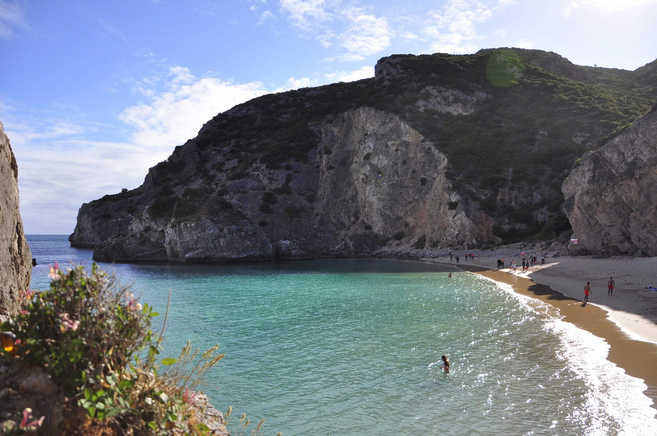 Beach scene with turquoise water, rocky cliffs, sandy shore, vegetation, and people swimming and walking