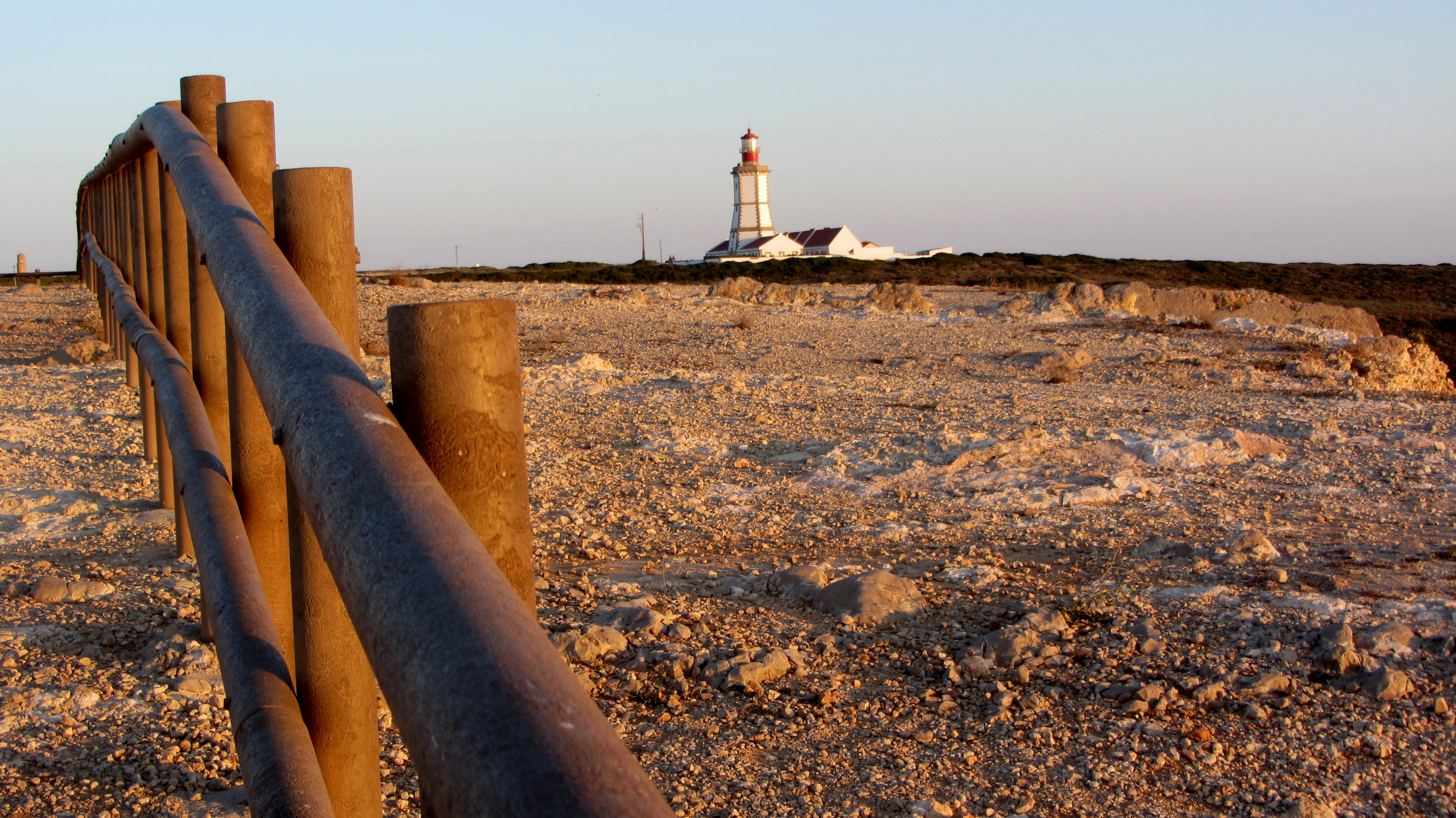 Rusty metal railing in foreground, rocky coastal terrain, and lighthouse with white and red stripes visible in the background under clear sky
