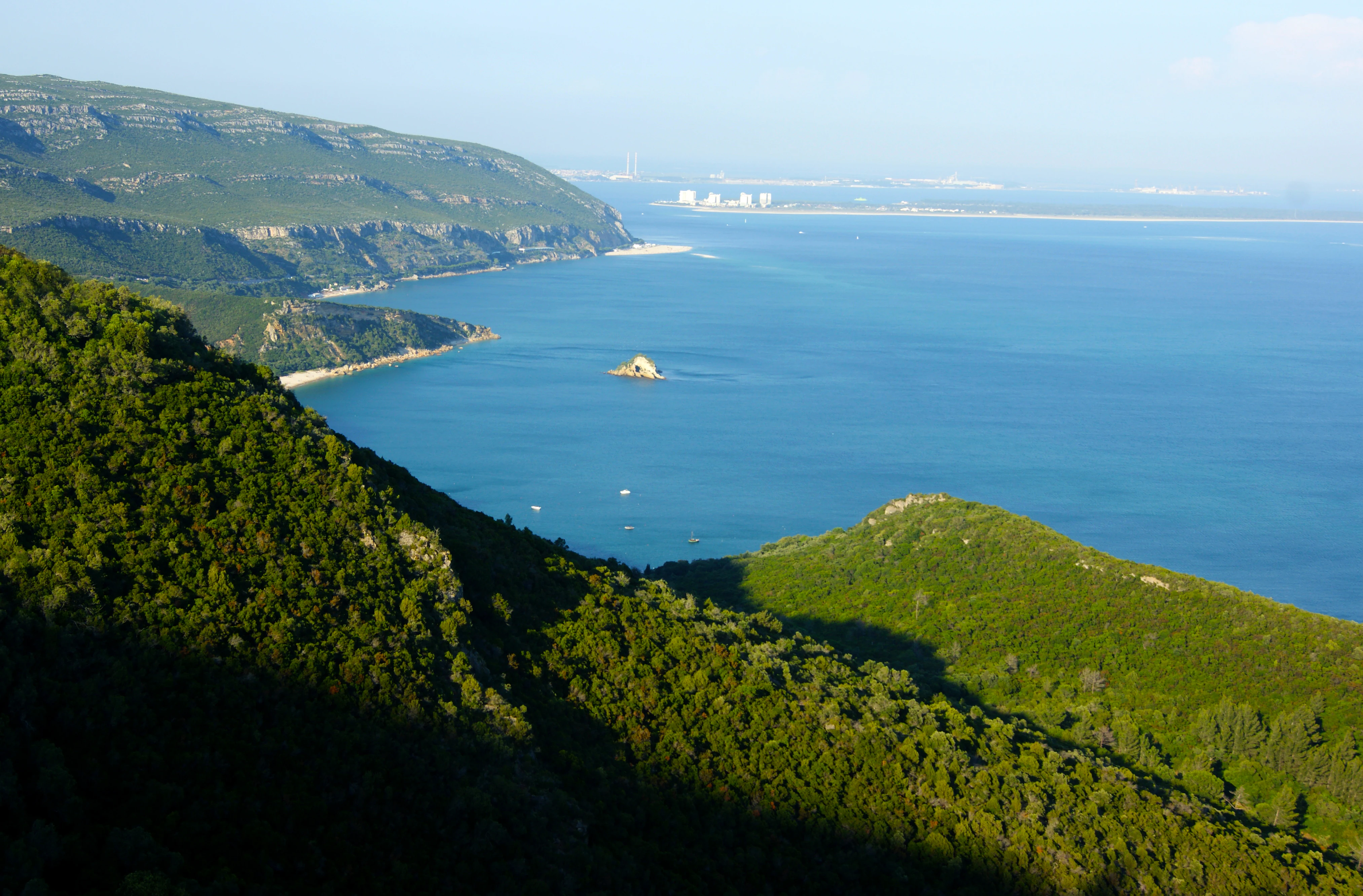 Green mountain slopes descending to a calm blue sea with a small rock island in the distance