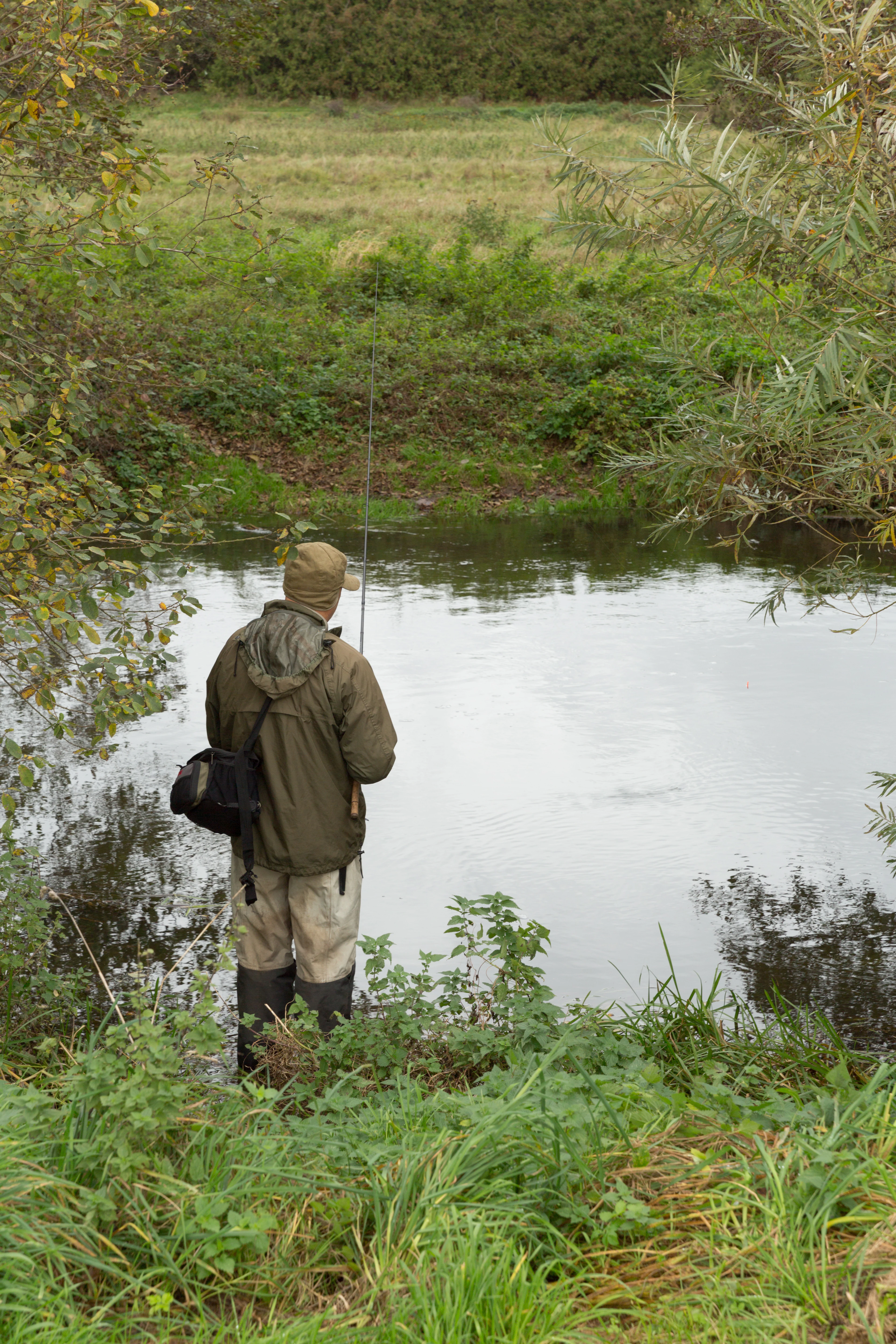Una persona con ropa de exterior y botas de agua está de pie en la orilla de un pequeño río tranquilo, sosteniendo una caña de pescar.