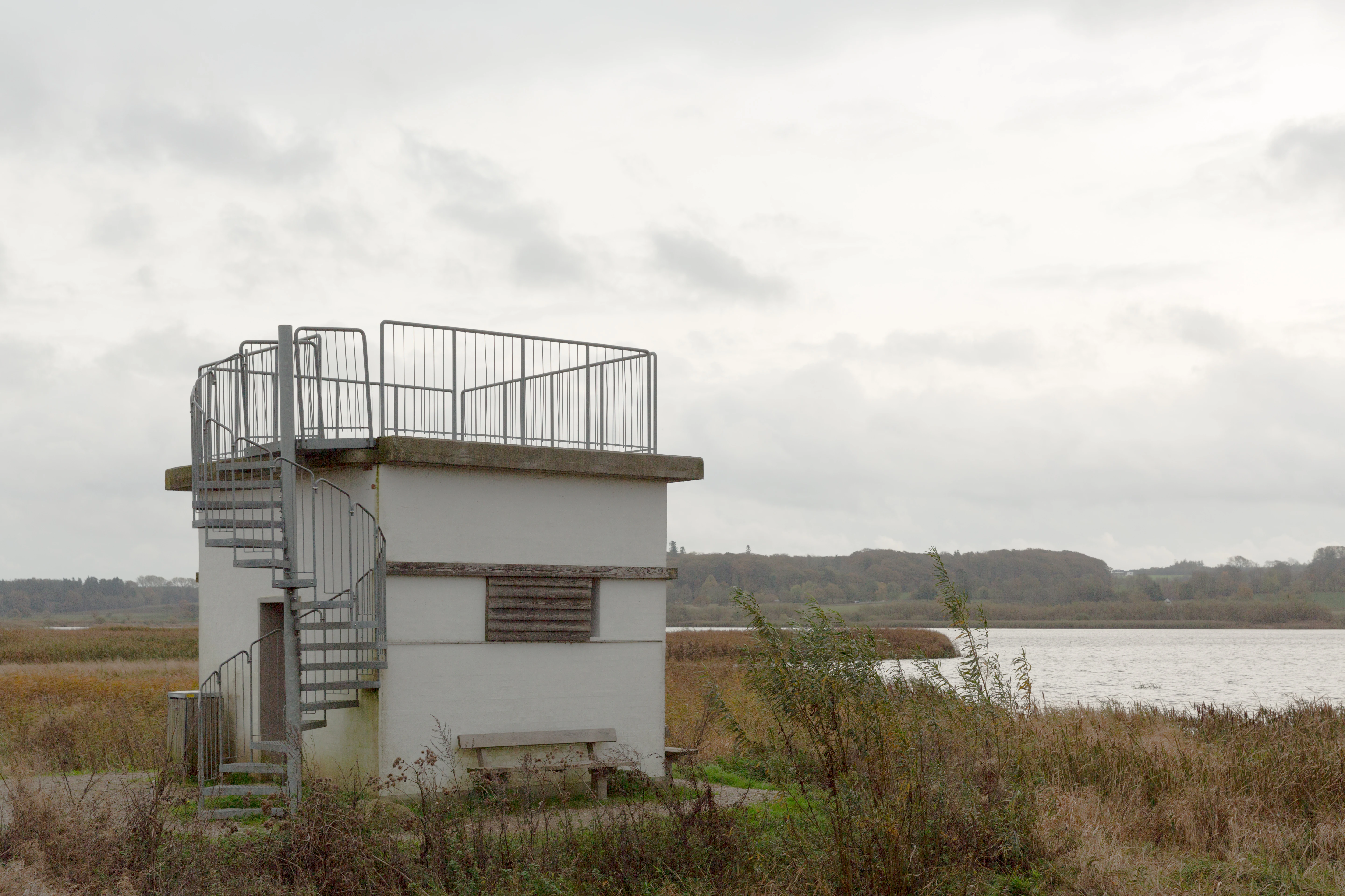 Una torre de observación de aves de hormigón blanco con una escalera metálica en espiral que conduce a una plataforma de observación al aire libre, situada en un paisaje de humedales con un lago al fondo bajo un cielo nublado.
