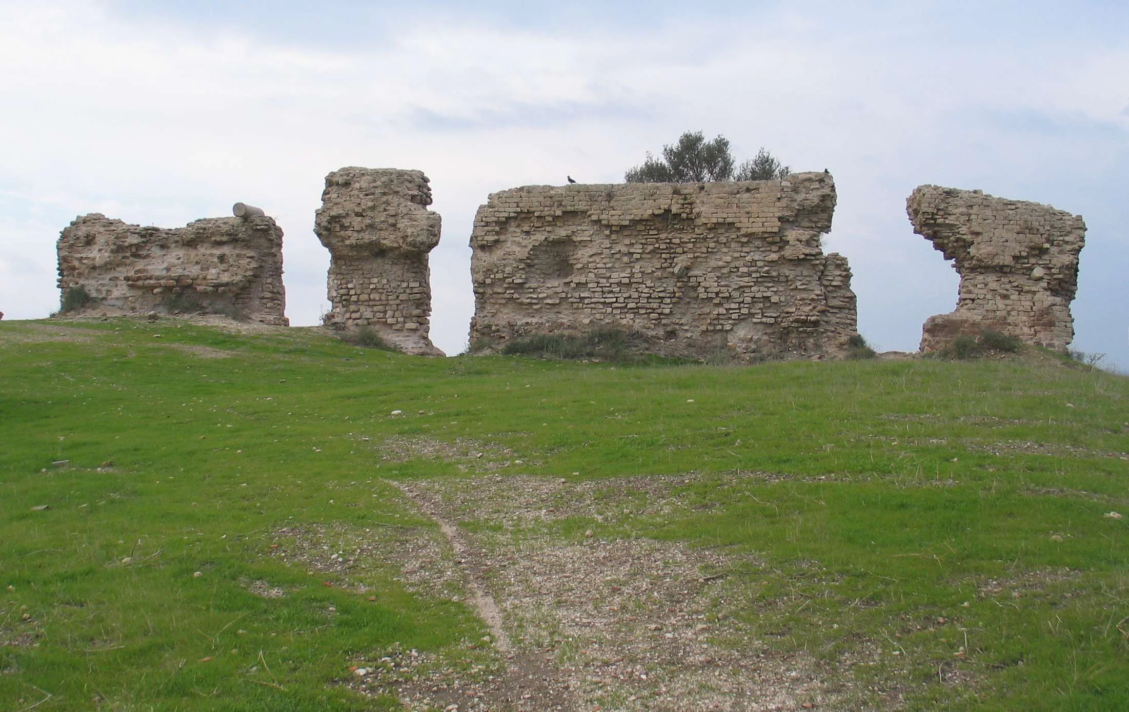 Stone ruins on a grassy hill with a dirt path leading up to them under a partly cloudy sky.