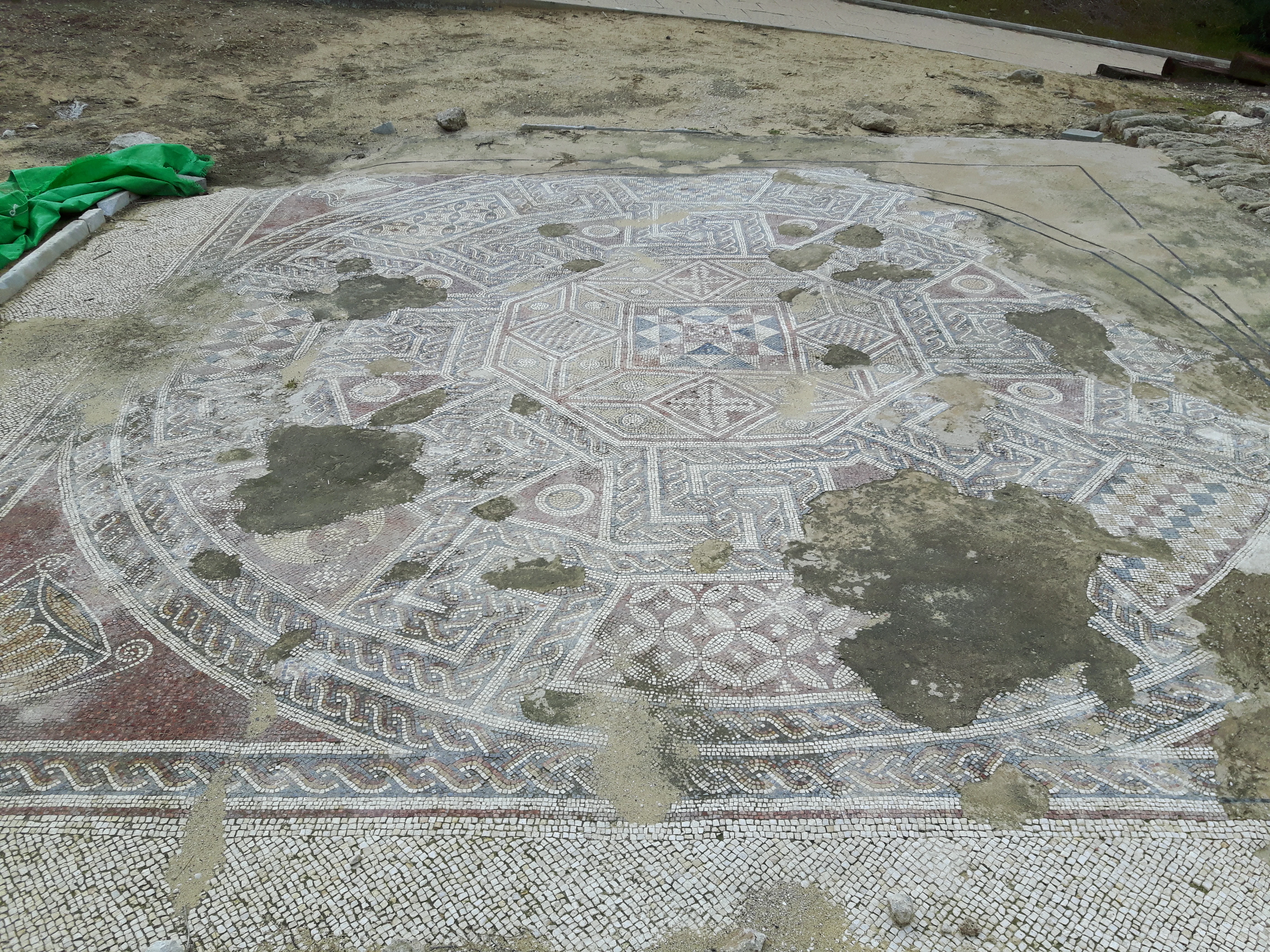 A large circular mosaic floor with geometric patterns and areas of missing tiles, situated in an archaeological site