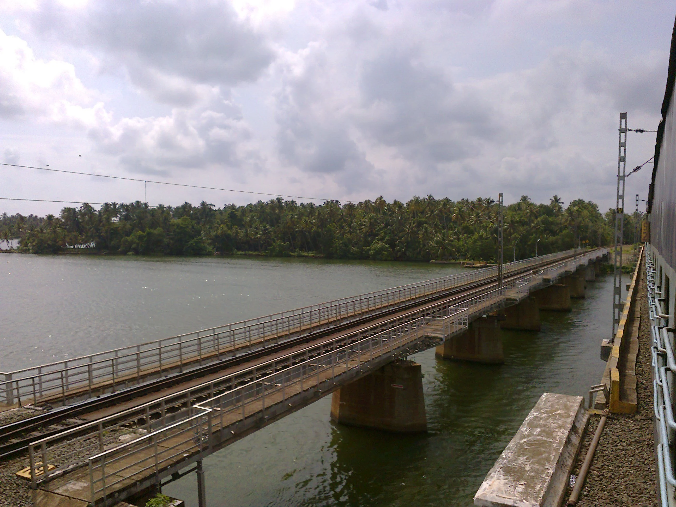 A view from a moving train looking across a railway bridge that spans the calm waters of Ashtamudi Lake, with lush green palm trees along the distant shoreline under a cloudy sky.