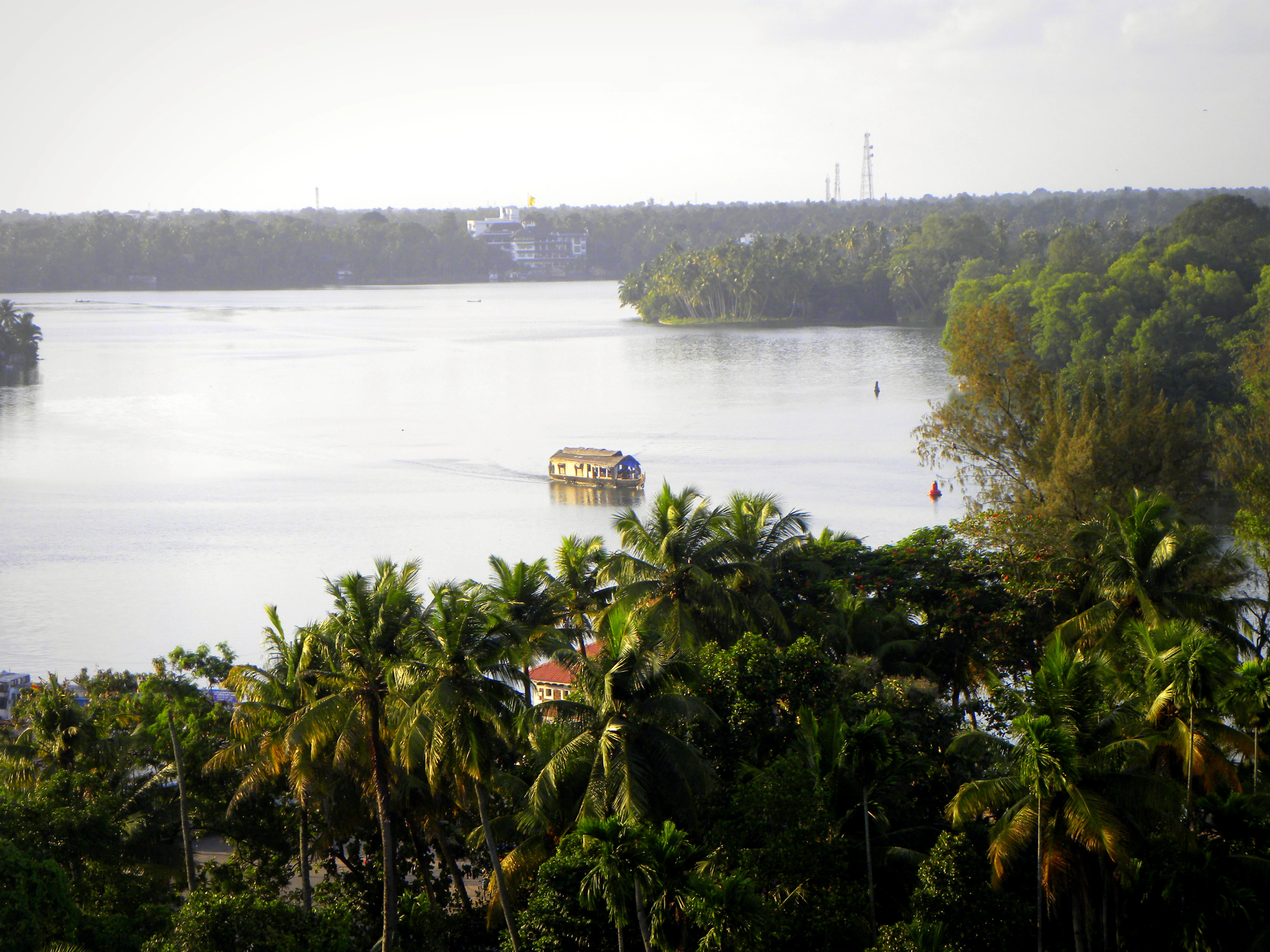 An elevated view of a wide lake in Kerala, India, featuring a traditional houseboat cruising through the center; palm trees line the shores.