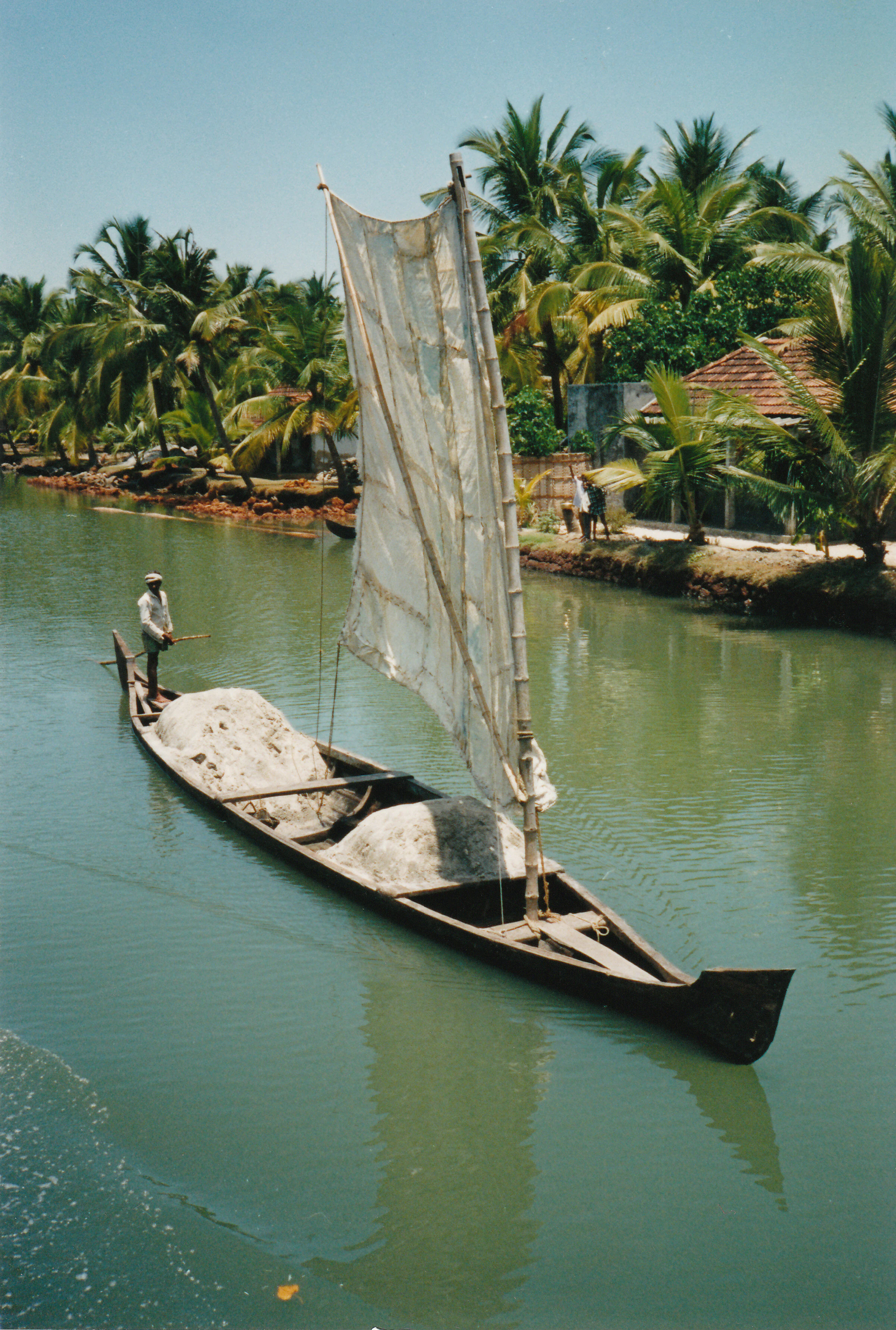A traditional narrow wooden boat with a white sail carrying piles of goods navigates a calm, narrow waterway lined with coconut palm trees in Kerala, India.