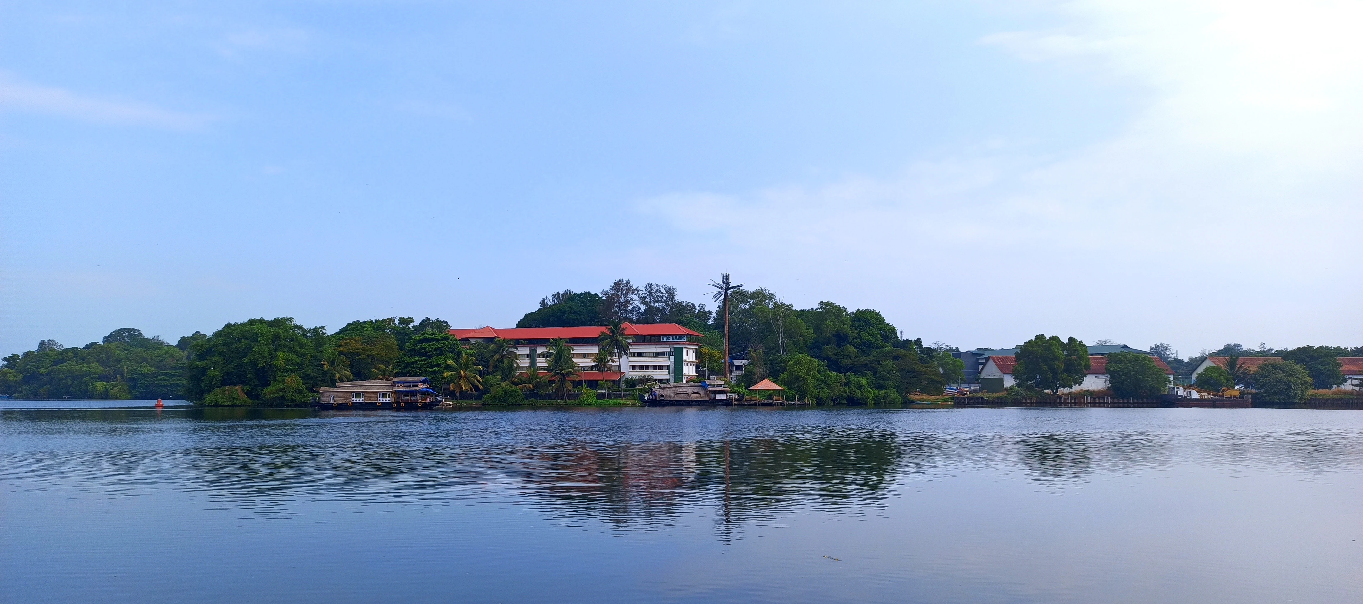 A wide shot of Ashtamudi Lake in Kerala featuring calm dark water reflecting a multi-story building and surrounding palm trees under a pale blue sky.