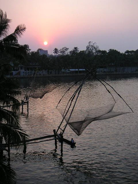 A sunset scene over calm water featuring traditional large wooden Chinese fishing nets, with a small boat nearby and palm trees on the distant shore.