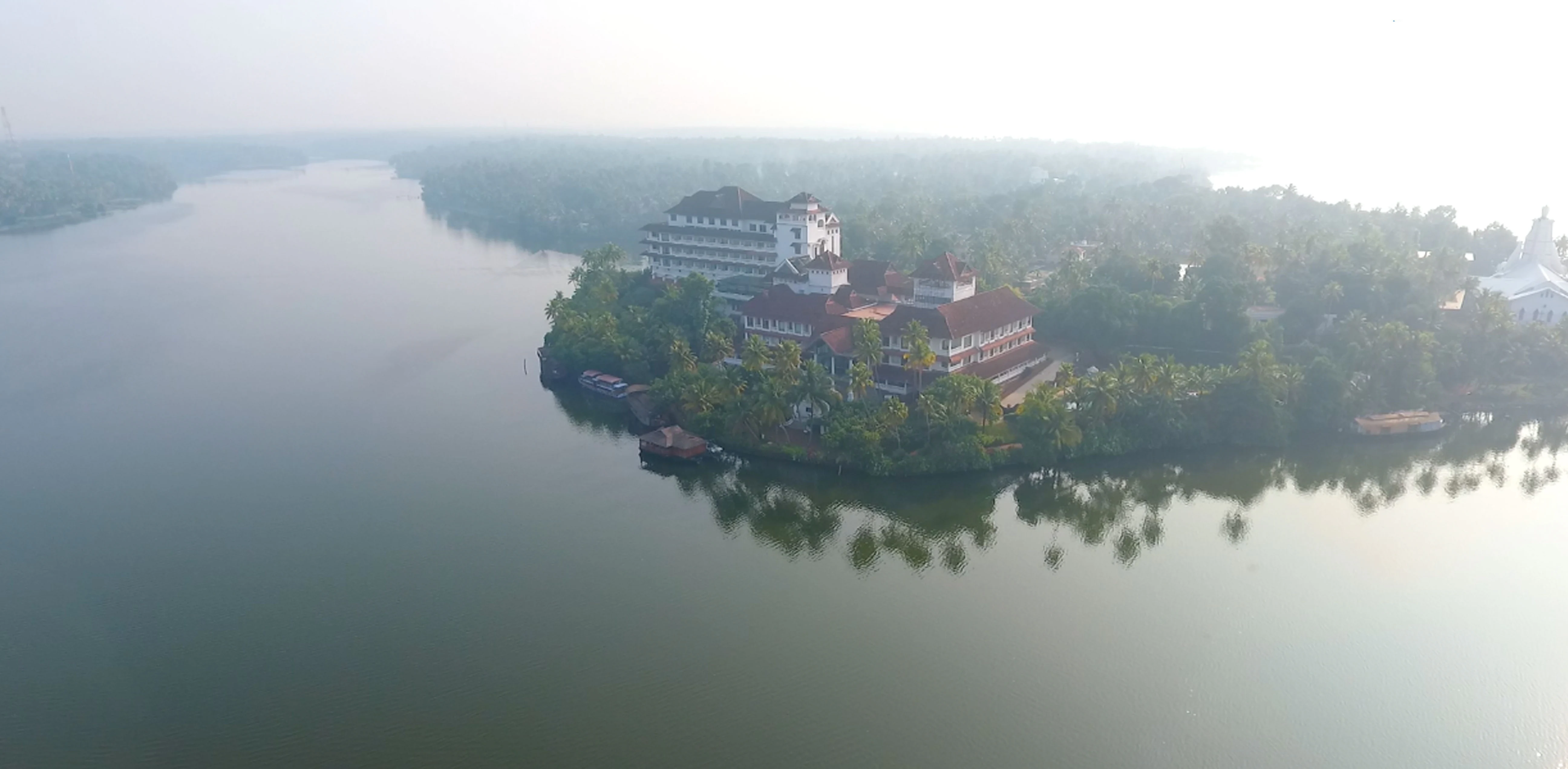 An aerial perspective of a large hotel complex situated on a lush, palm-covered peninsula surrounded by the calm, hazy waters of Ashtamudi Lake.