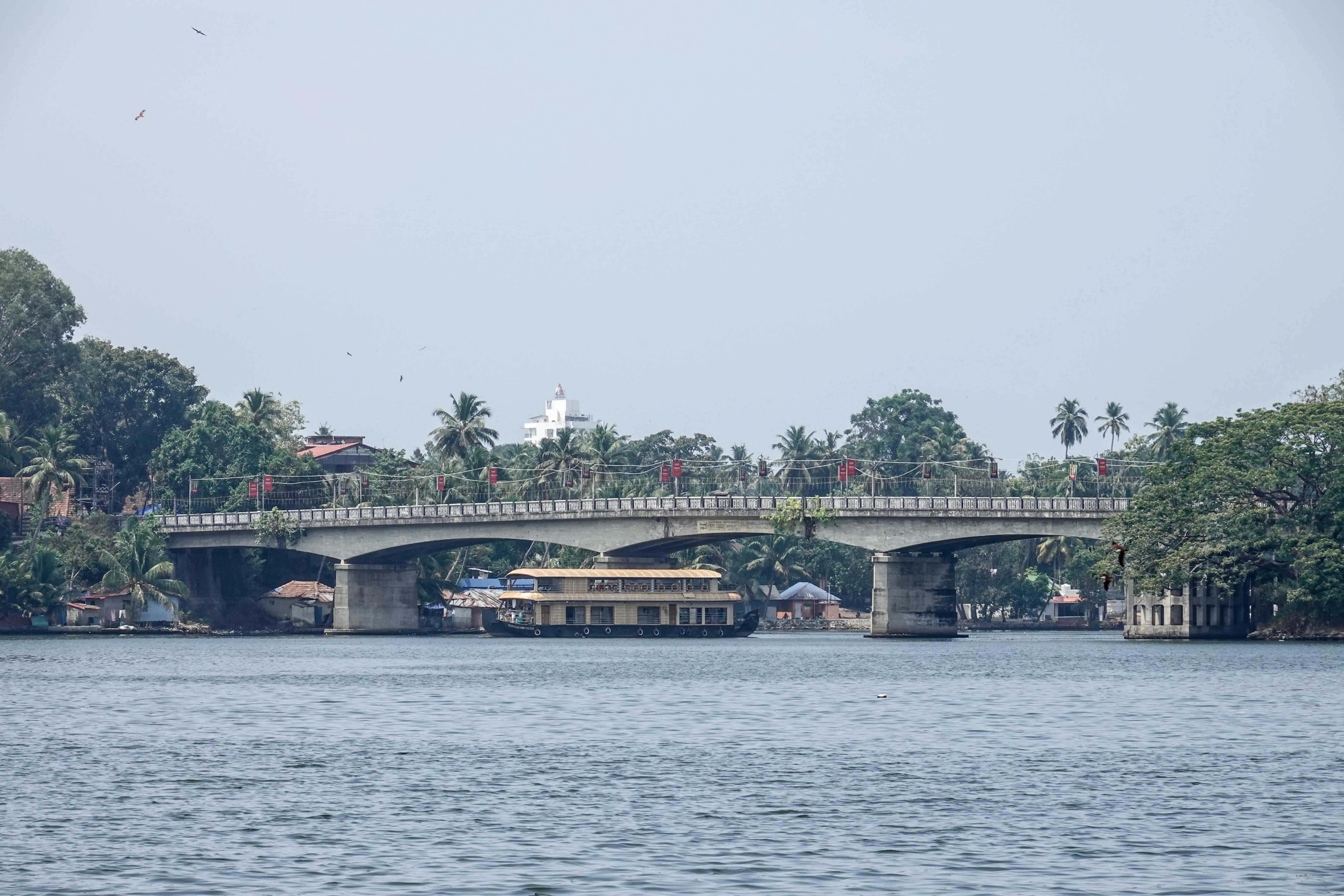 A traditional wooden houseboat cruises under a concrete bridge spanning a wide lake, surrounded by lush green trees.