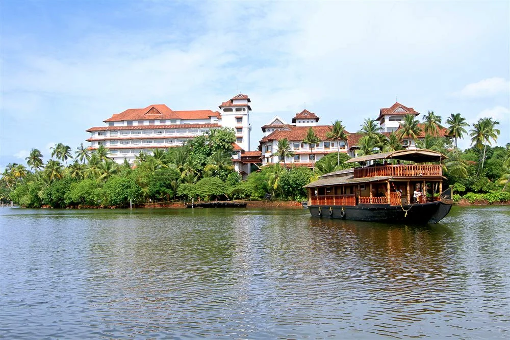 A traditional wooden houseboat floating on the calm waters of Ashtamudi Lake with a large resort building and dense palm trees on the shoreline.
