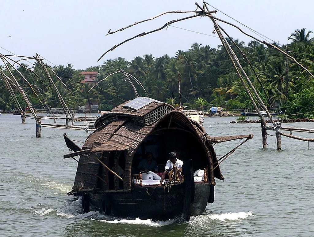 A traditional houseboat with a thatched roof cruising on the calm waters of Ashtamudi Lake in Kerala, with several large Chinese fishing nets visible in the background against a backdrop of palm trees.